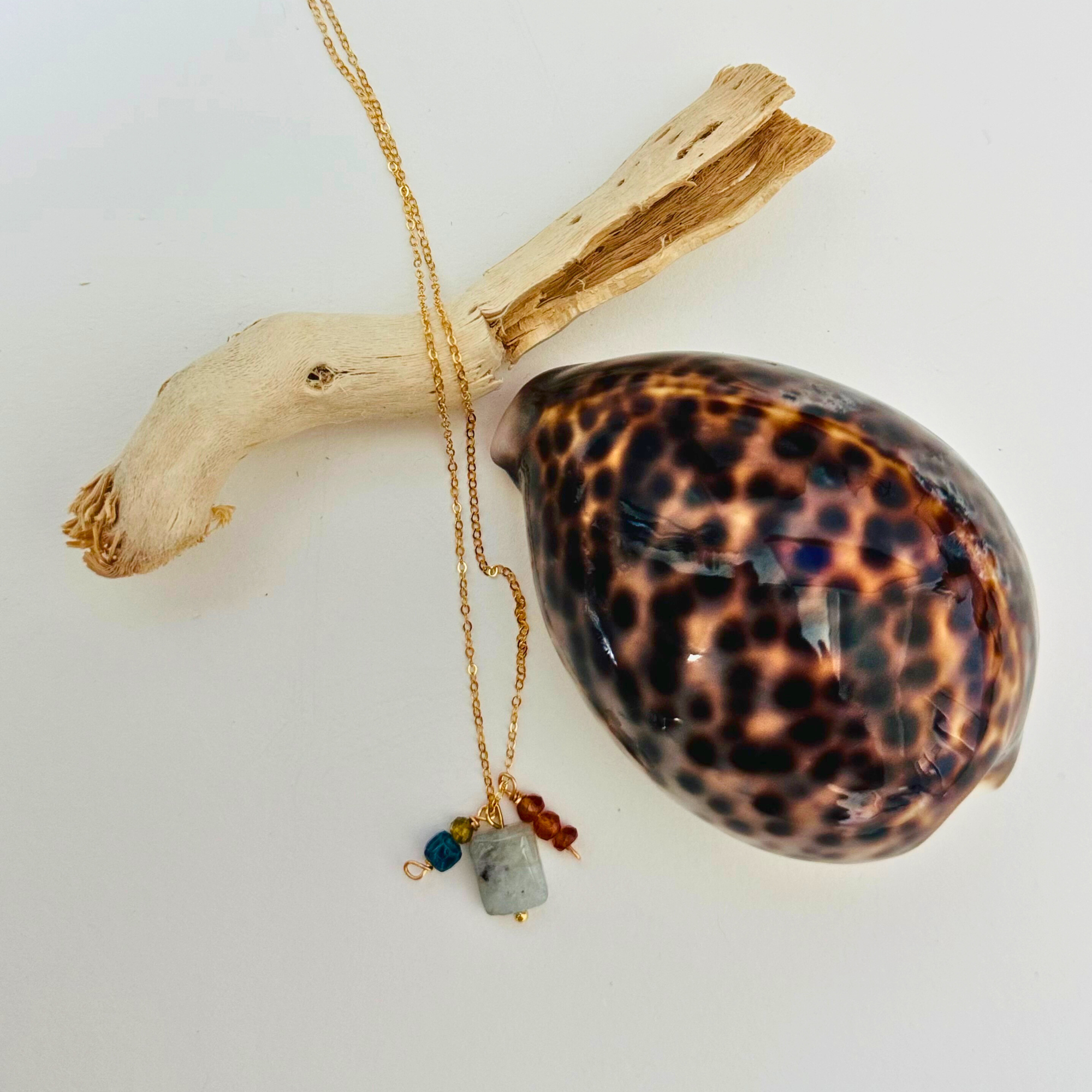 A dainty gold necklace with aventurine, mystic blue topaz, citrine, and garnet hessonite stones is draped over a bleached driftwood branch and framed by a glossy seashell.