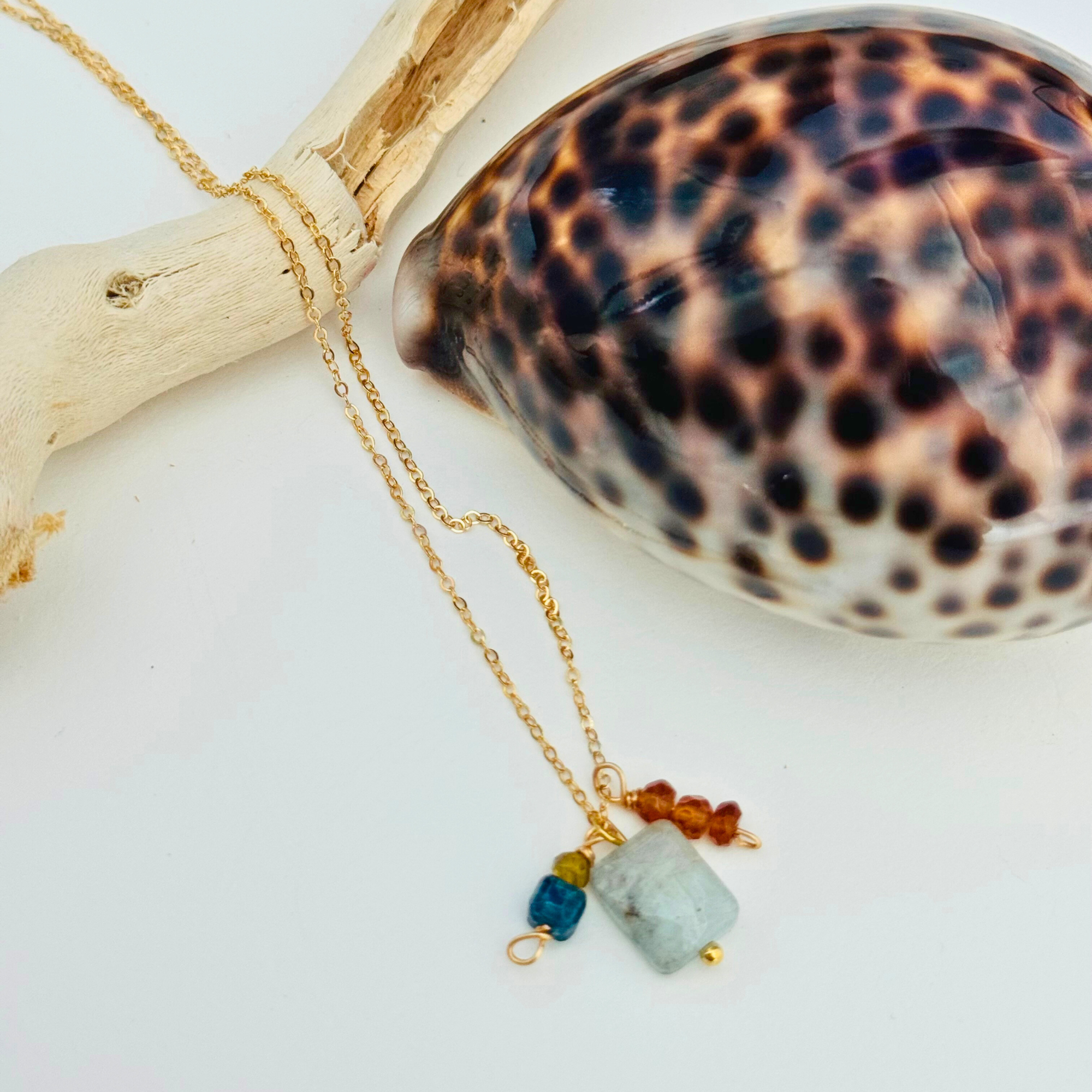 A dainty gold necklace with aventurine, mystic blue topaz, citrine, and garnet hessonite stones is draped over a bleached driftwood branch and framed by a glossy seashell.