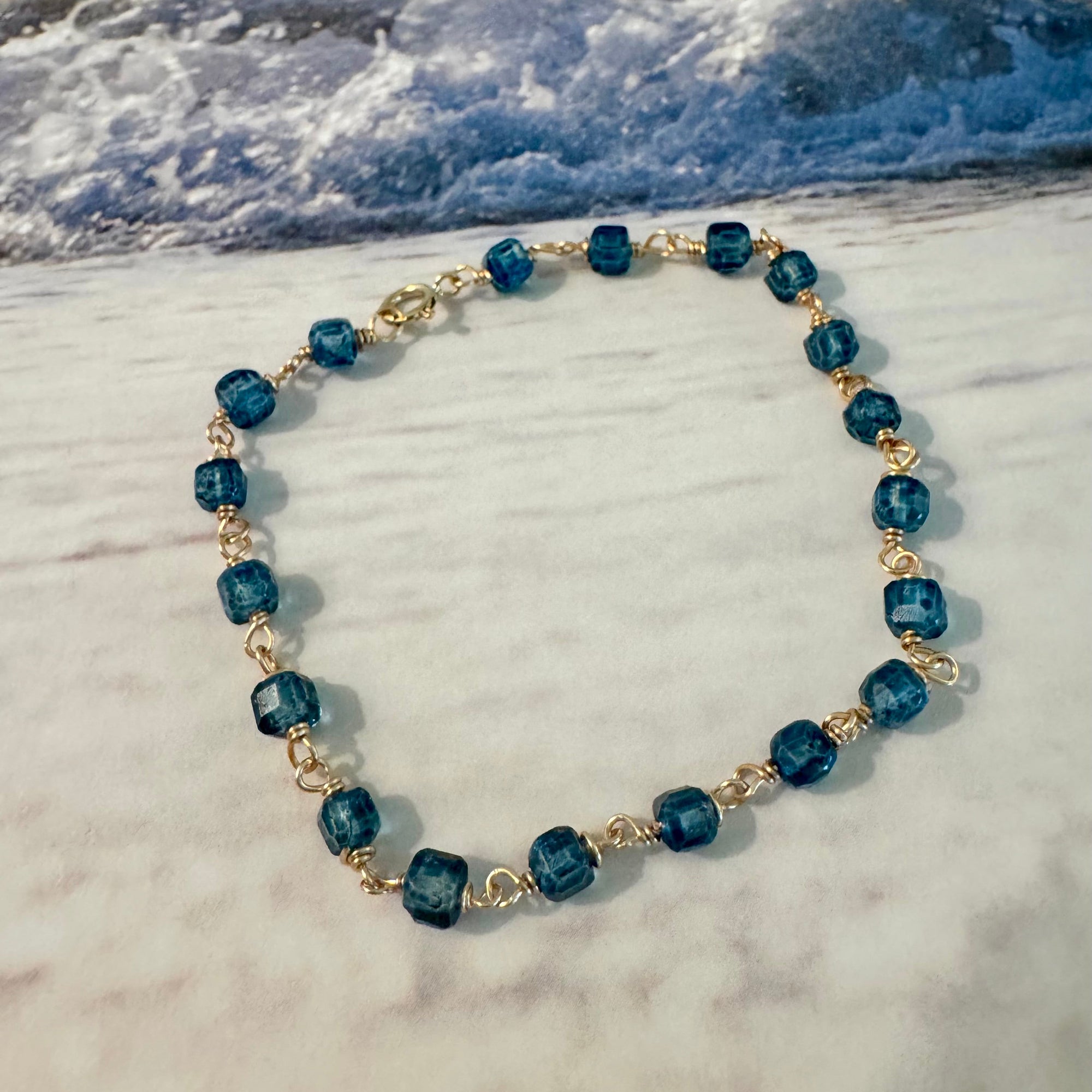 Blue Seas Topaz Bracelet made with cube-faceted blue topaz beads wrapped in 14k gold filled wire is displayed against a blue and white beach surf background.