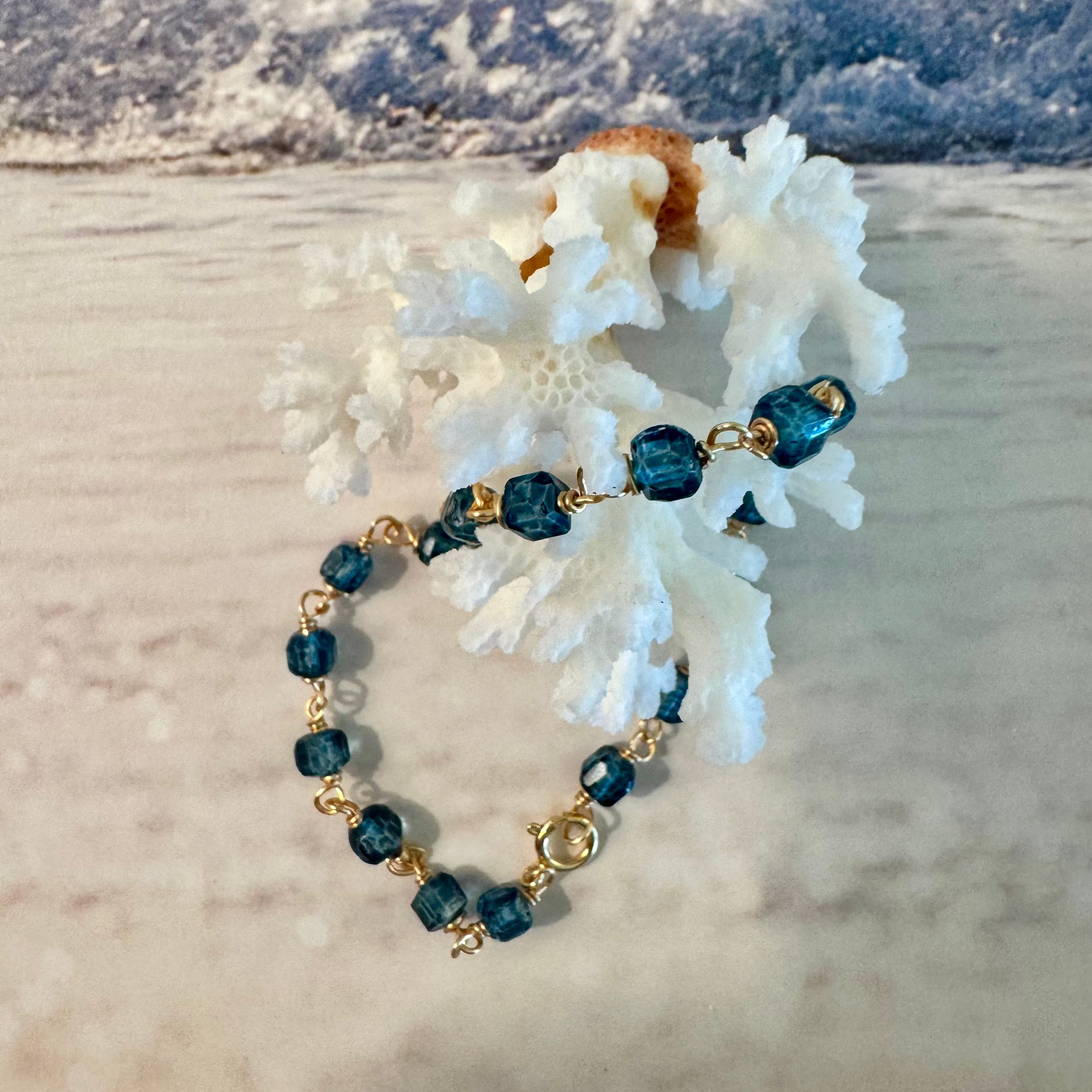 Blue Seas Topaz Bracelet made with cube-faceted blue topaz beads wrapped in 14k gold filled wire is displayed twisted around white coral against a blue and white beach surf background.