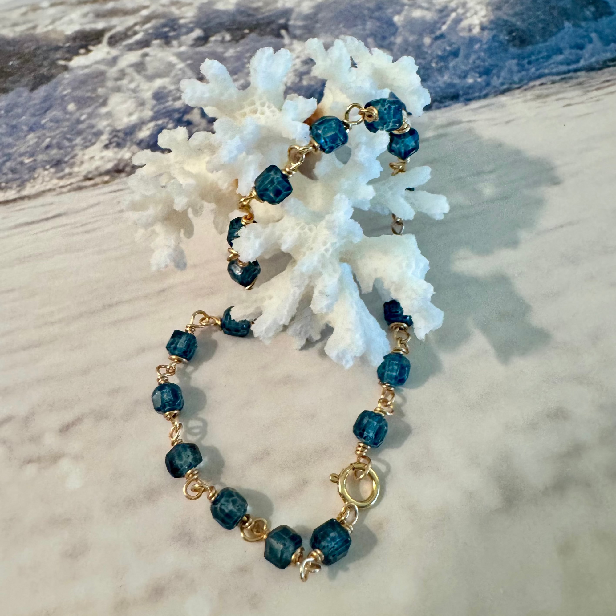 Blue Seas Topaz Bracelet made with cube-faceted blue topaz beads wrapped in 14k gold filled wire is displayed twisted around white coral against a blue and white beach surf background at a different angle showing the clasp closure.