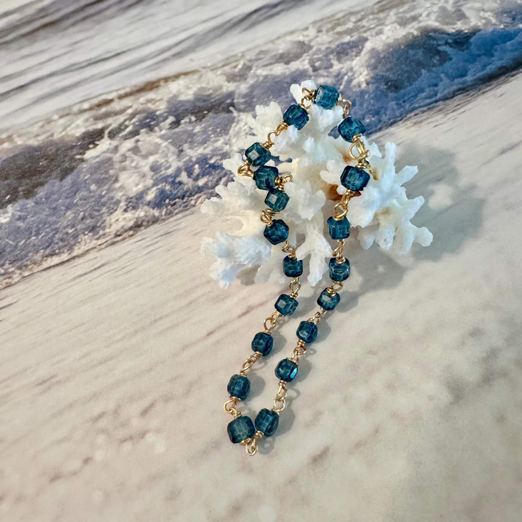 Blue Seas Topaz Bracelet made with cube-faceted blue topaz beads wrapped in 14k gold filled wire is displayed draped over white coral against a blue and white beach surf background.