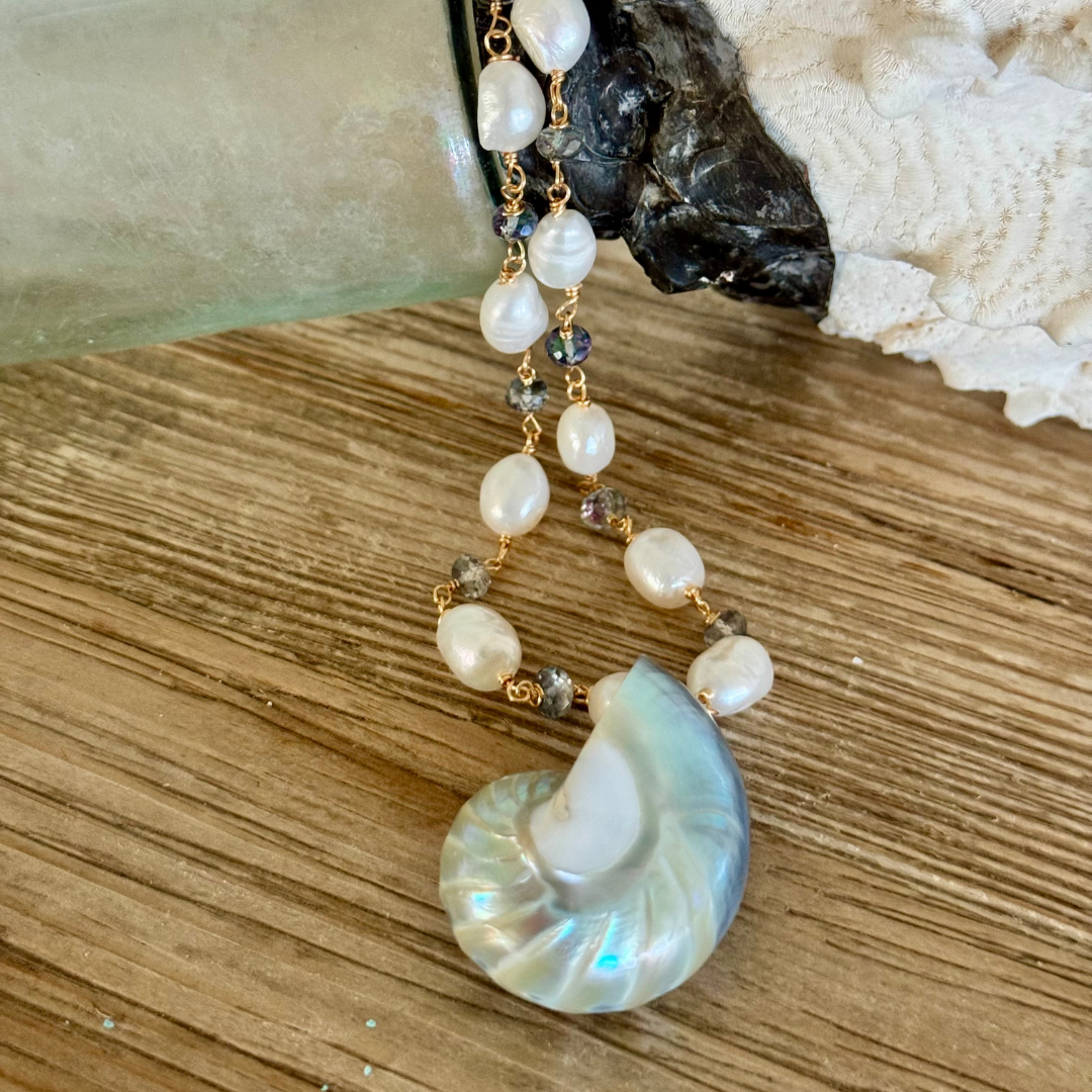 Genuine blue nautilus shell pendant draped over coral jar display on beachy wood surface. Freshwater pearls and mystic topaz accents between 14k gold-filled wire and chain.
