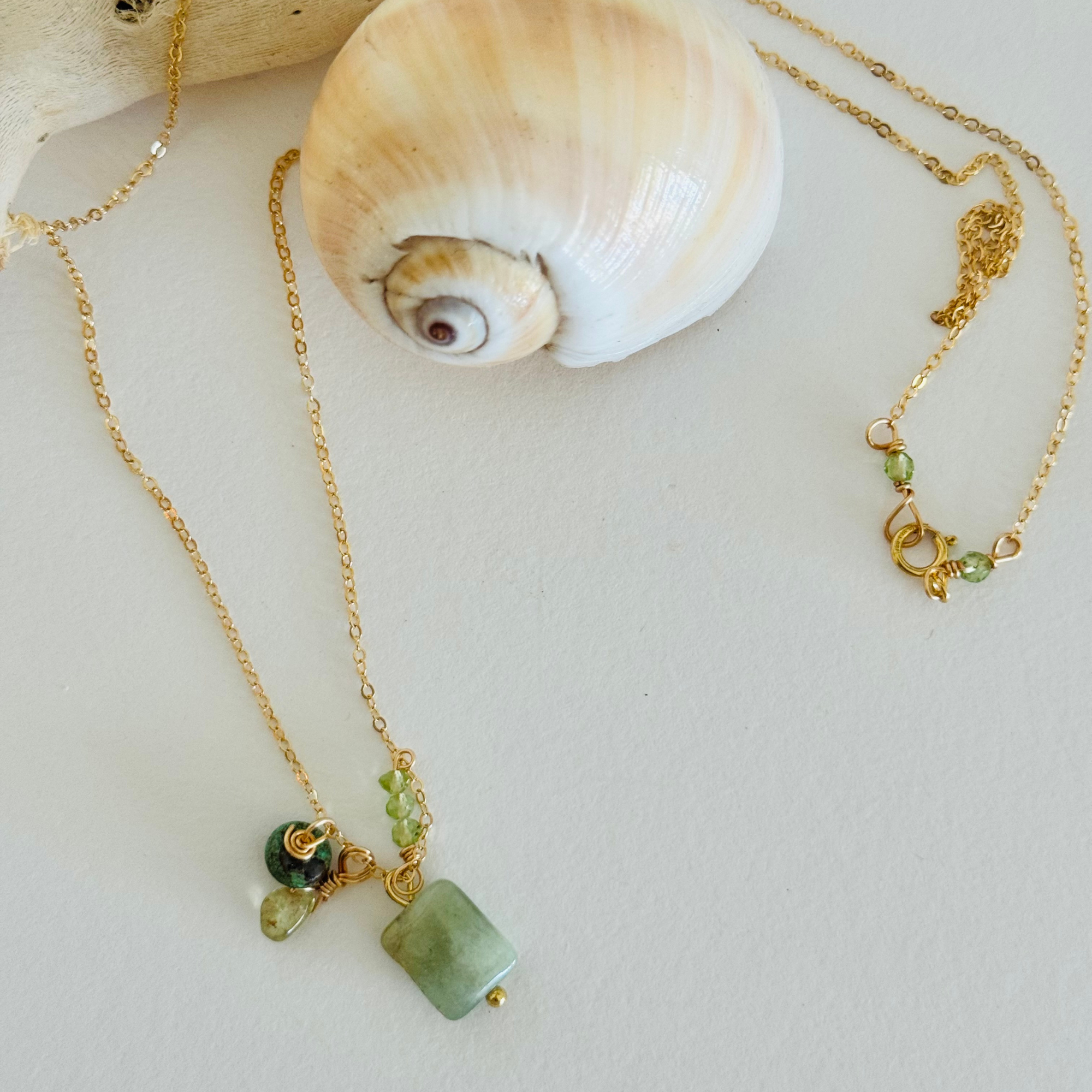A necklace is draped over a creamy spiral seashell and bleached driftwood. It is made of a simple gold chain with aventurine, peridot, and turquoise. A pair of small light green beads offset the clasp.