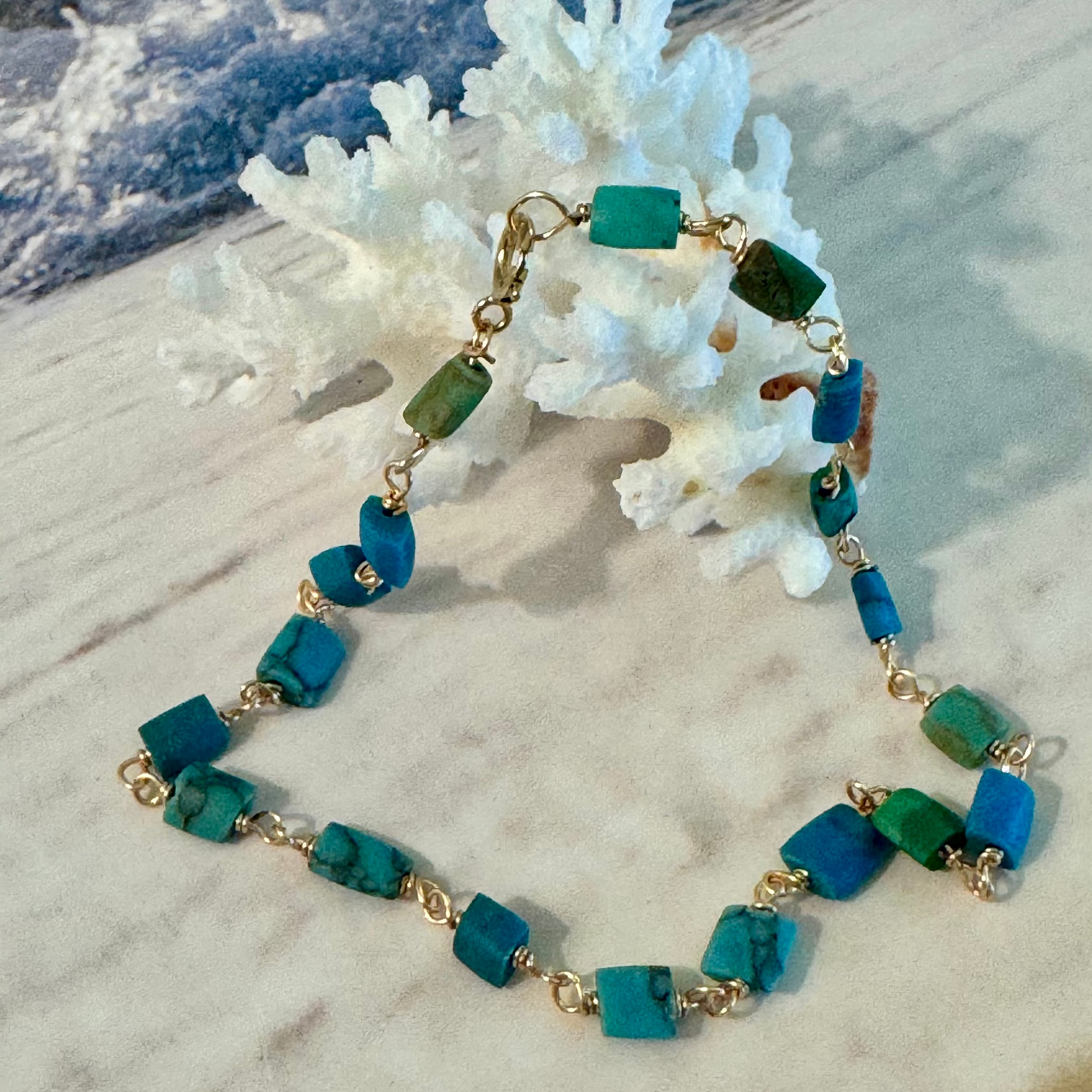 A bracelet made of genuine matte turquoise beads with a smooth, flat varying in intensity of green to blue on 14k gold filled wire. Displayed against white coral on a beachy background.