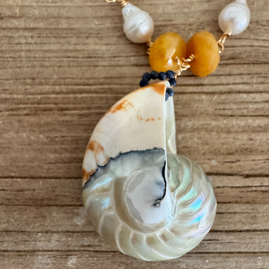 Closeup view of genuine nautilus shell pendant with iridescent shimmer, showcasing navy blue and pumpkin orange veining in the shell. Small navy beads and a pair of yellow-orange beads flank the shell, then teardrop pearls offset them and wrap up around the rest of the 14k gold filled wire chain, displayed against a beachy wood grain surface.