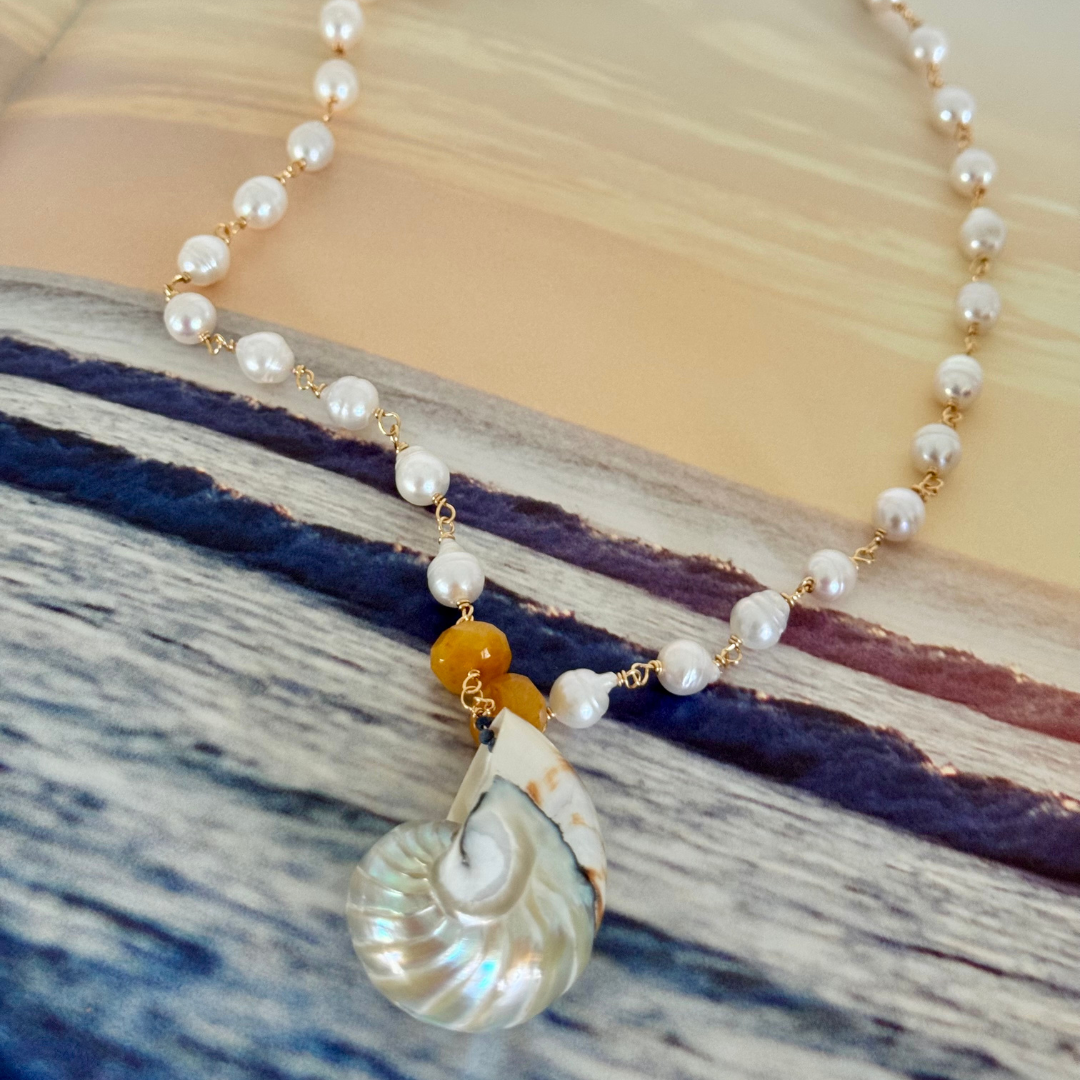 Nautilus shell pendant displayed against a magazine page showcasing navy ocean waves set against a yellow clouded sky at sunrise or sunset, viewed at 45-degree angle