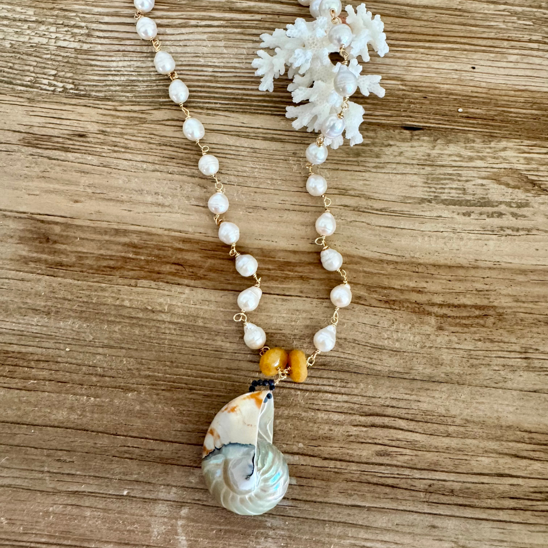 Nautilus shell pendant displayed on white coral and against beachy wood grain surface.
