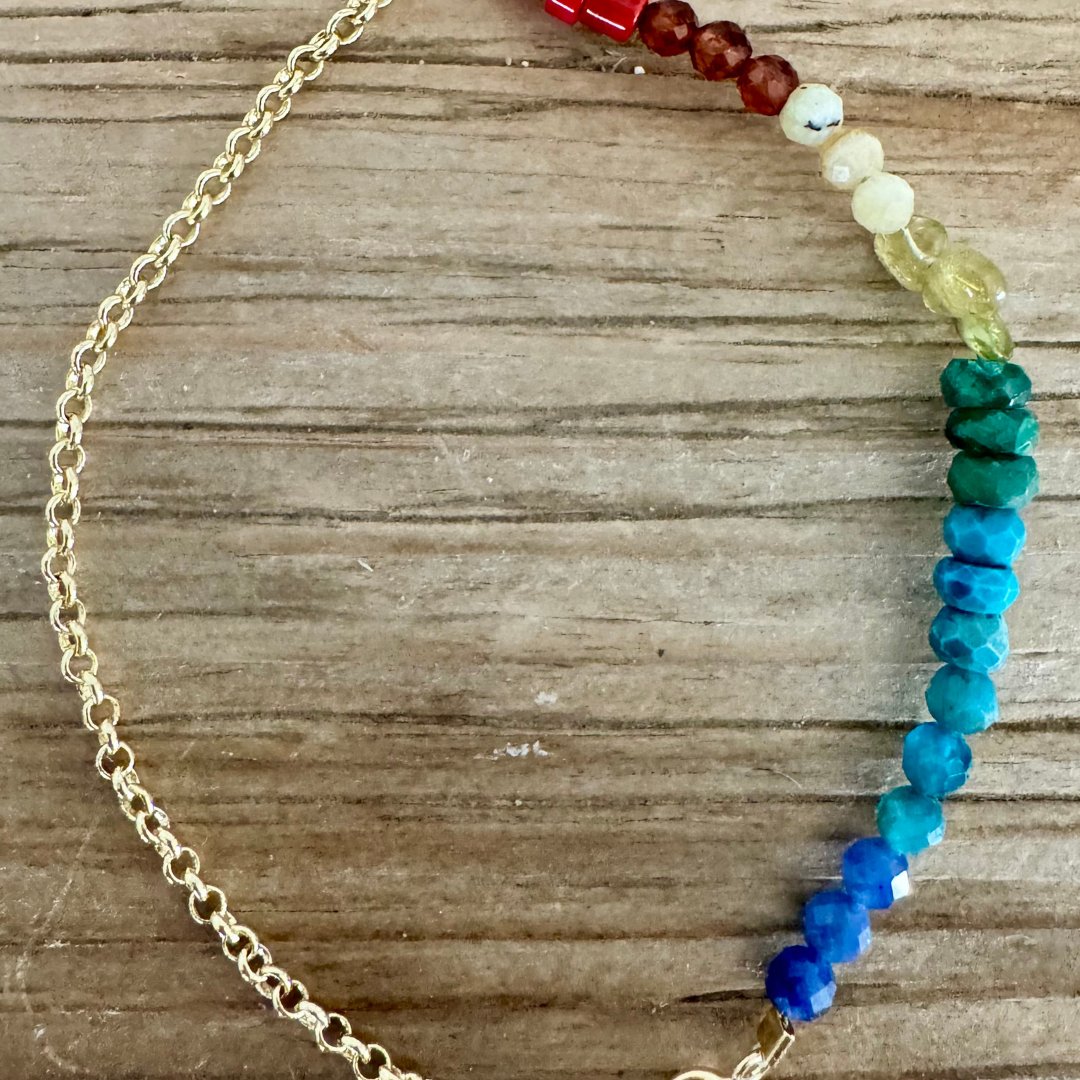 A bracelet is displayed on woodgrain surface. The bracelet is 14k gold chain with a pearl at the clasp. There are beads on half of the bracelet in three of each color of bright red, scarlet orange, milky yellow, glassy yellow-green, emerald green, blue, turquoise, and cerulean. Stunning rainbow ombre effect.