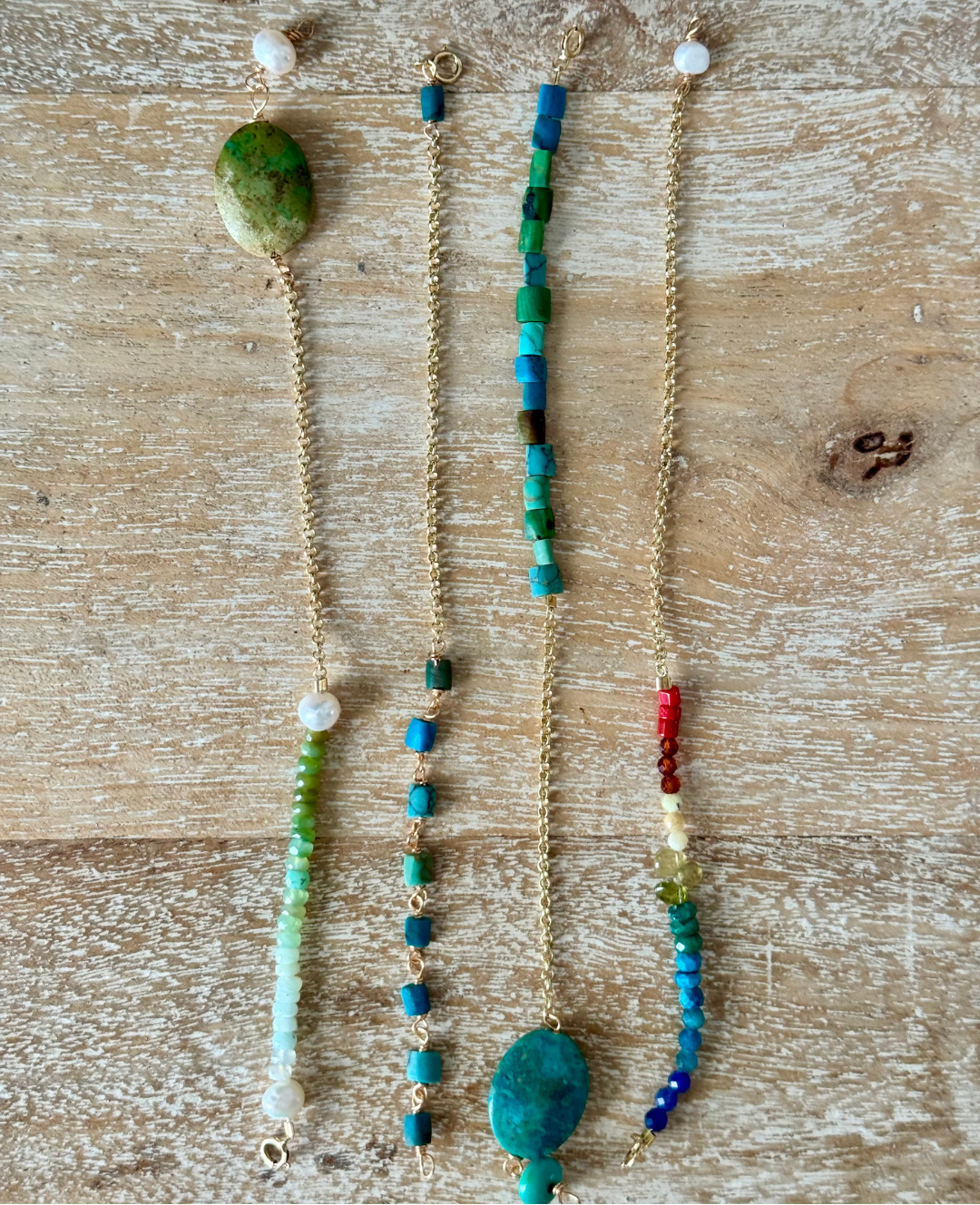 A set of four bracelets are laid out on a beachy wooden surface from an aerial view. From left to right is a green seafoam opal pearl bracelet, blue and green square bead bracelet, turquoise opal bracelet, and beaded rainbow dreams bracelet, all with 14k gold chains. Creates a potential stack of bracelets.