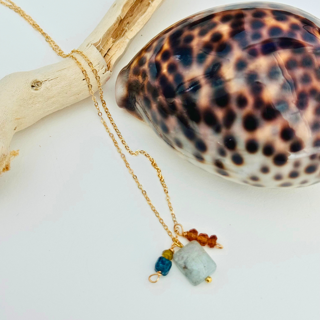 A dainty gold necklace with aventurine, mystic blue topaz, citrine, and garnet hessonite stones is draped over a bleached driftwood branch and framed by a glossy seashell.