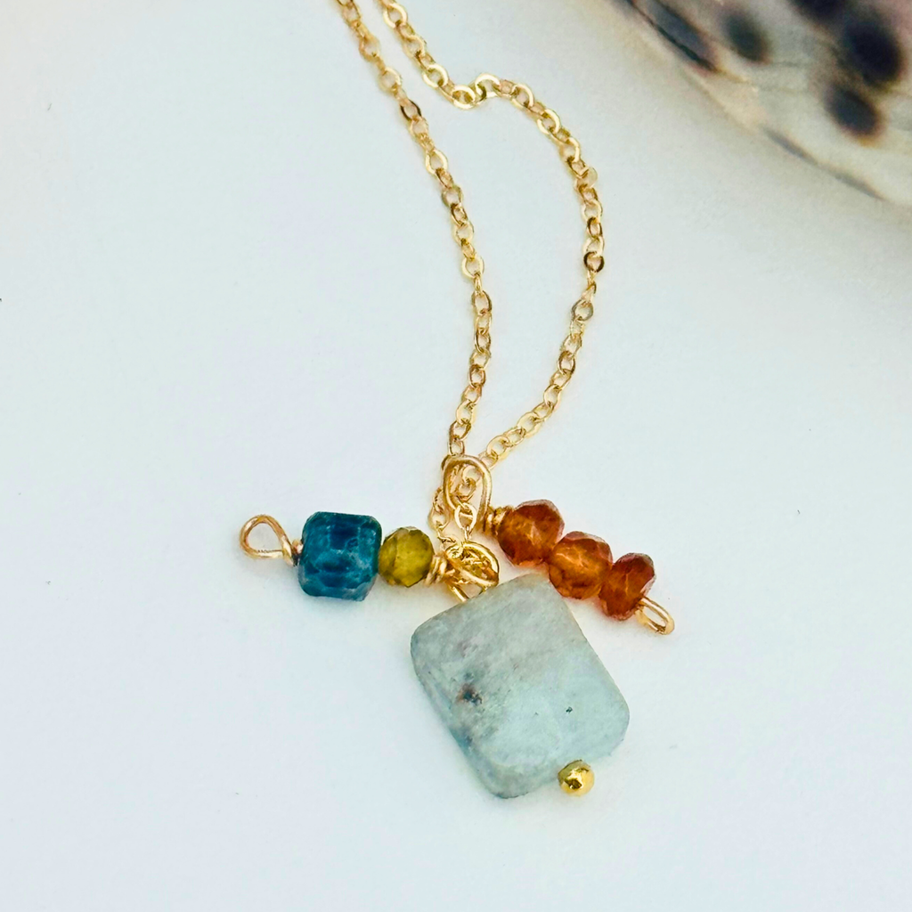 Closeup of a dainty gold necklace with aventurine, mystic blue topaz, citrine, and garnet hessonite stones on white background.
