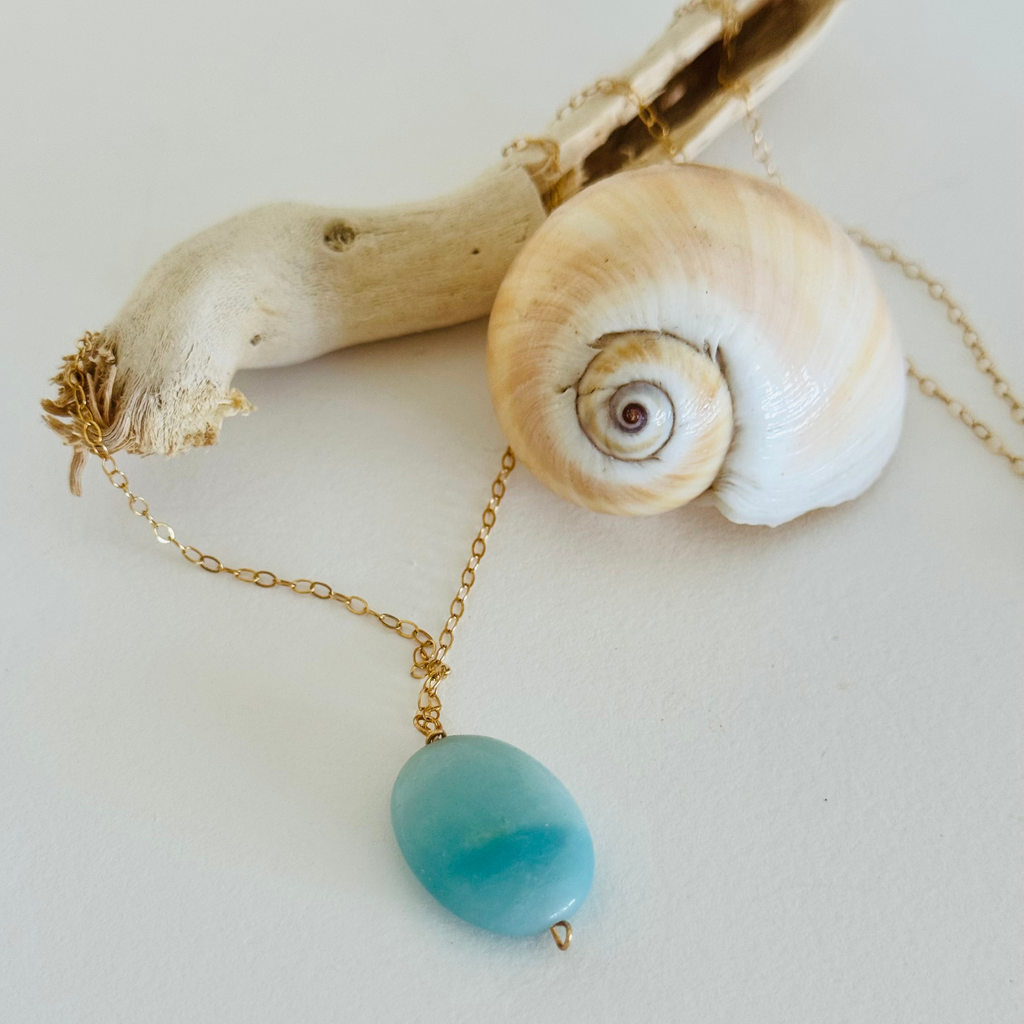 Aquamarine sea drop pendant with light blue color variation on 14k gold filled wire chain draped across a spiral seashell and bleached driftwood branch.