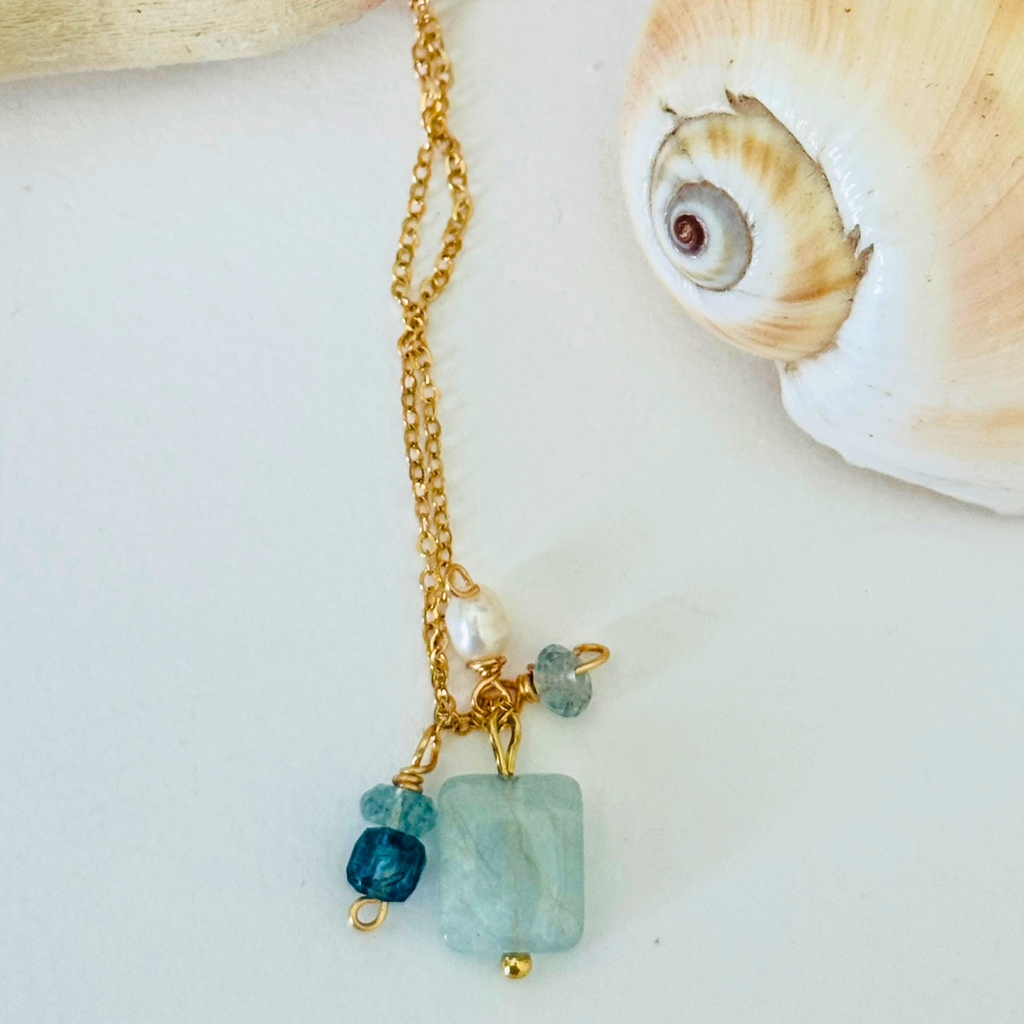 Close up view of a simple dainty 14k gold filled wire chain necklace made of aventurine, mystic blue topaz, aquamarine, and freshwater pearl stones. There are five stones total ranging from white pearl to light blue to navy blue. Displayed on a white background, framed by a creamy white spiral seashell.