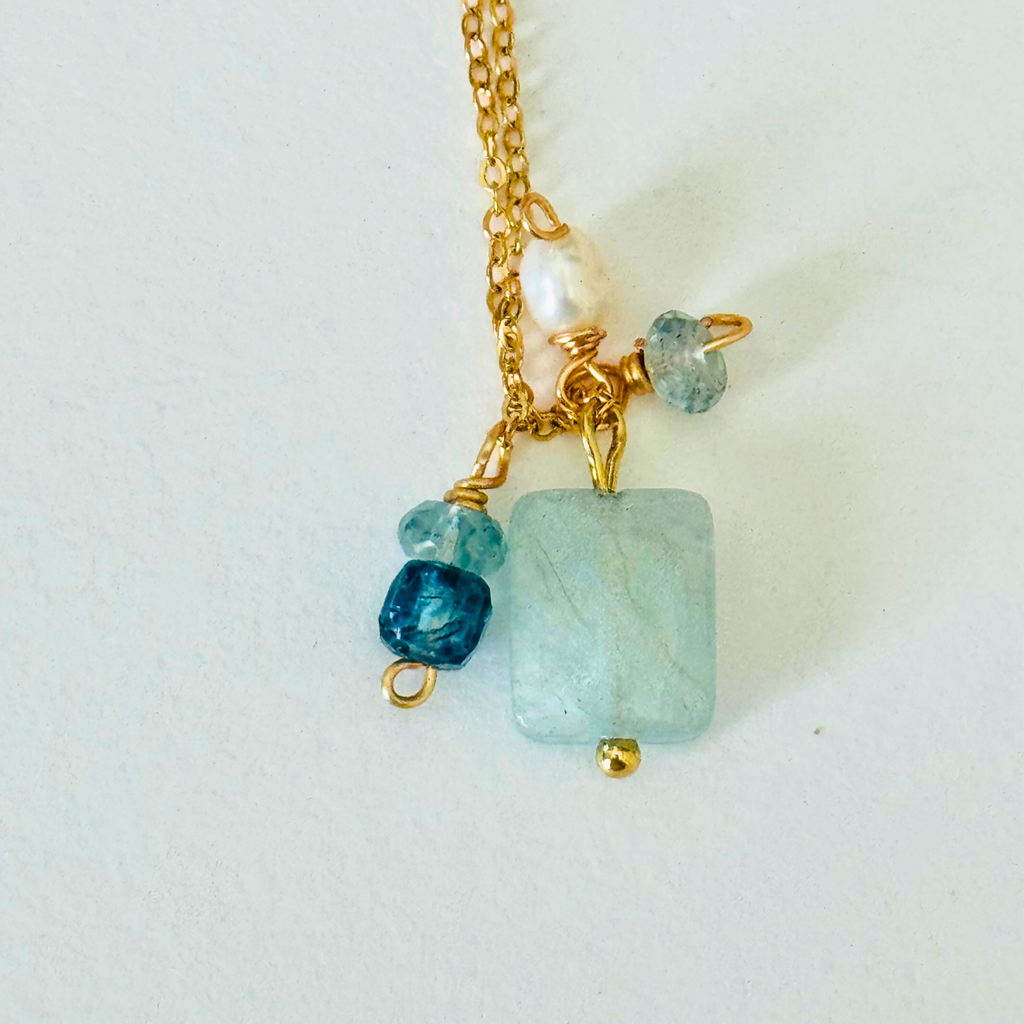 Close up view of a simple dainty 14k gold filled wire chain necklace made of aventurine, mystic blue topaz, aquamarine, and freshwater pearl stones. There are five stones total ranging from white pearl to light blue to navy blue. Displayed on a white background.