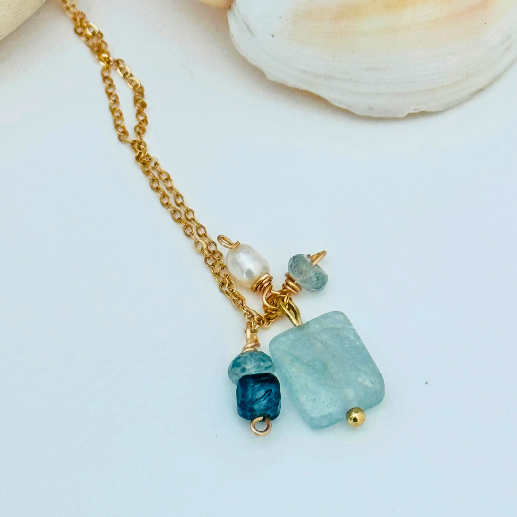 Close up view of a simple dainty 14k gold filled wire chain necklace made of aventurine, mystic blue topaz, aquamarine, and freshwater pearl stones. There are five stones total ranging from white pearl to light blue to navy blue. Displayed at an angle on a white background beneath a white seashell.