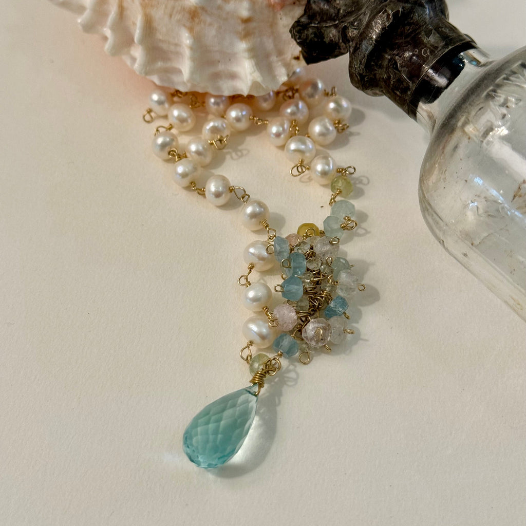 Pearl necklace with a blue teardrop pendant on a light surface with a shell and bottle.