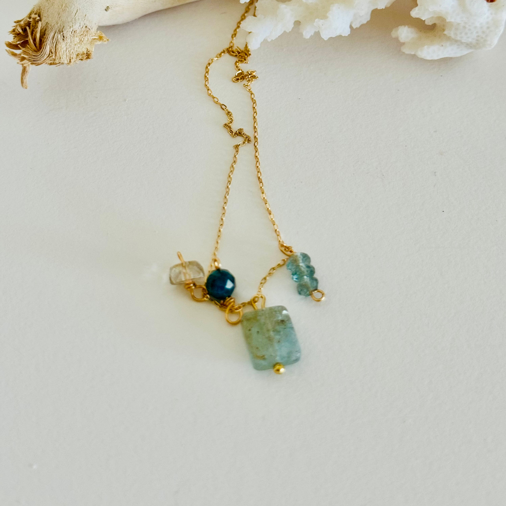 Aventurine, Apatite, Mystic Topaz, and Gold Quartz stones (one of each) on 14k gold-filled wire chain. Blue and green hues in the sones and translucent so the wiring can ever so slightly be seen through, creating contrast and warmth. Whole chain extended long, framed by white coral and bleached driftwood. On white background.