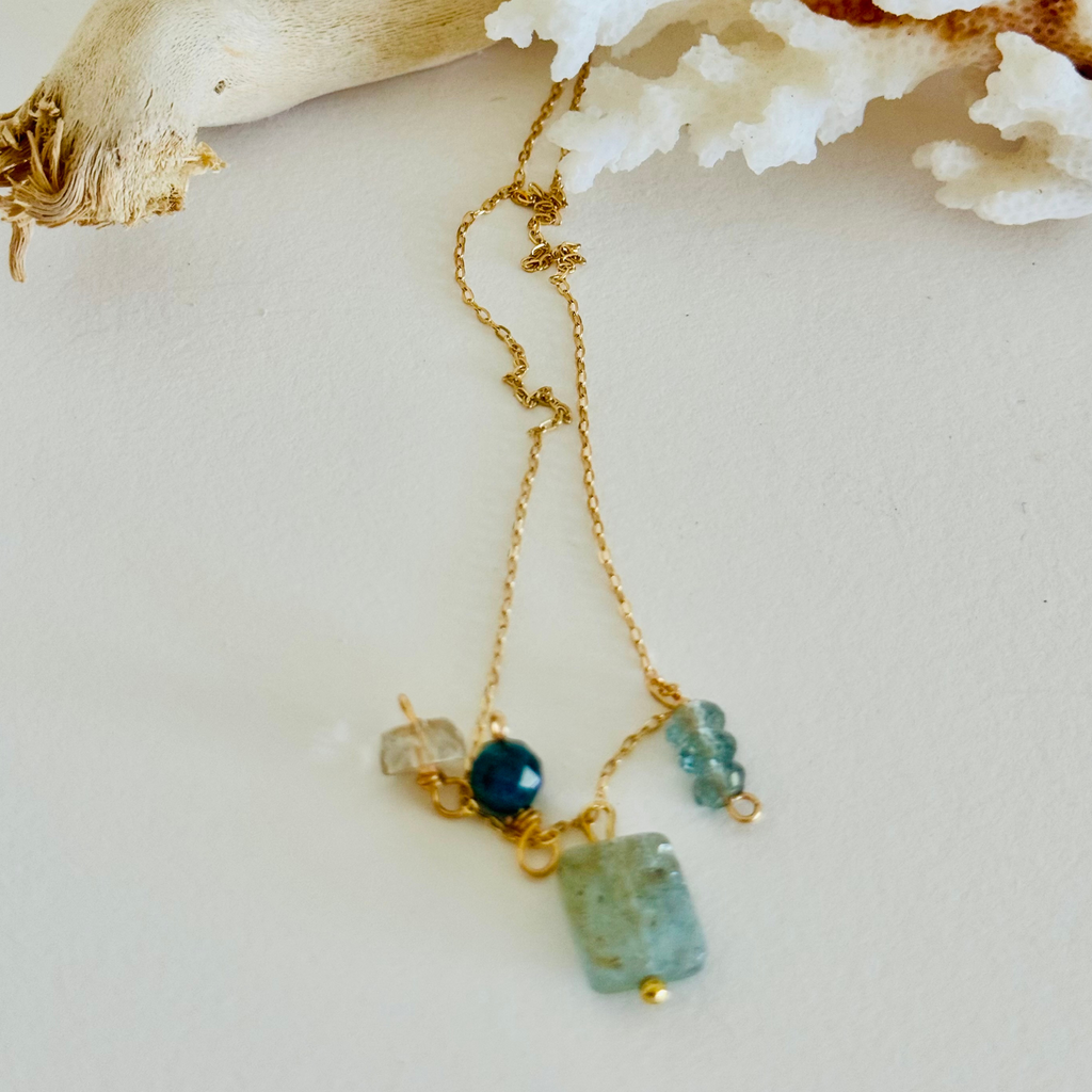 Aventurine, Apatite, Mystic Topaz, and Gold Quartz stones (one of each) on 14k gold-filled wire chain. Blue and green hues in the sones and translucent so the wiring can ever so slightly be seen through, creating contrast and warmth. Whole chain extended long, framed by white coral and bleached driftwood. On white background.