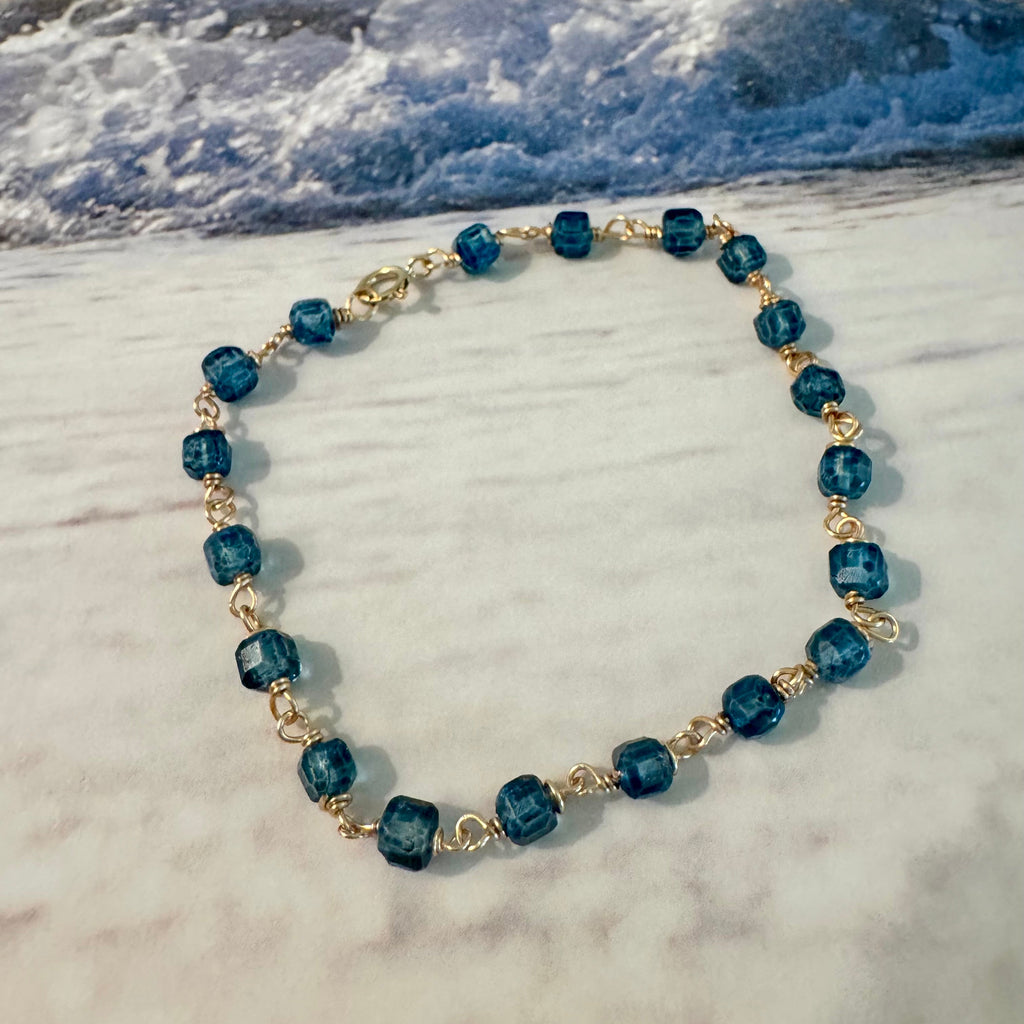 Blue Seas Topaz Bracelet made with cube-faceted blue topaz beads wrapped in 14k gold filled wire is displayed against a blue and white beach surf background.