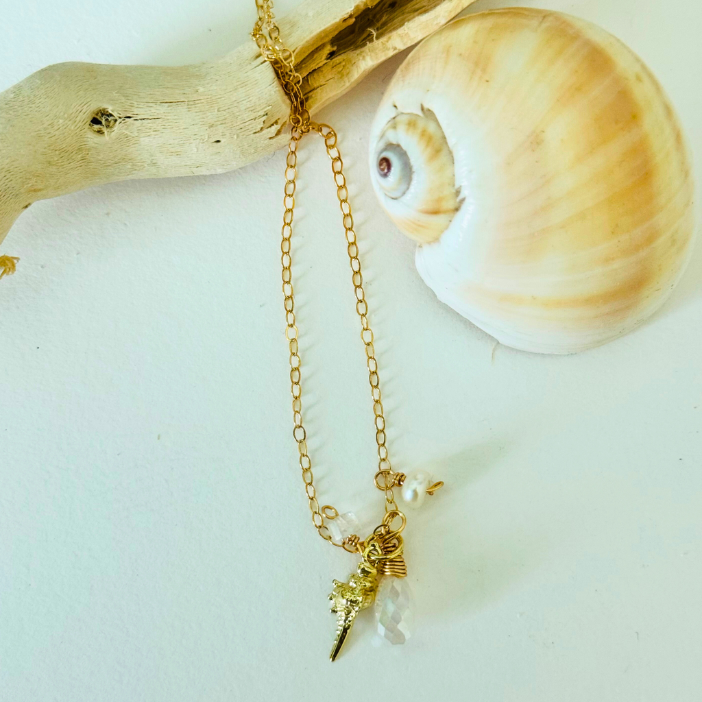 A simple dainty 14k gold filled wire necklace with pearl, aquamarine, and moonstone stones and a gold seashell charm are draped over bleached driftwood and framed by a creamy warm spiral seashell on a white background.