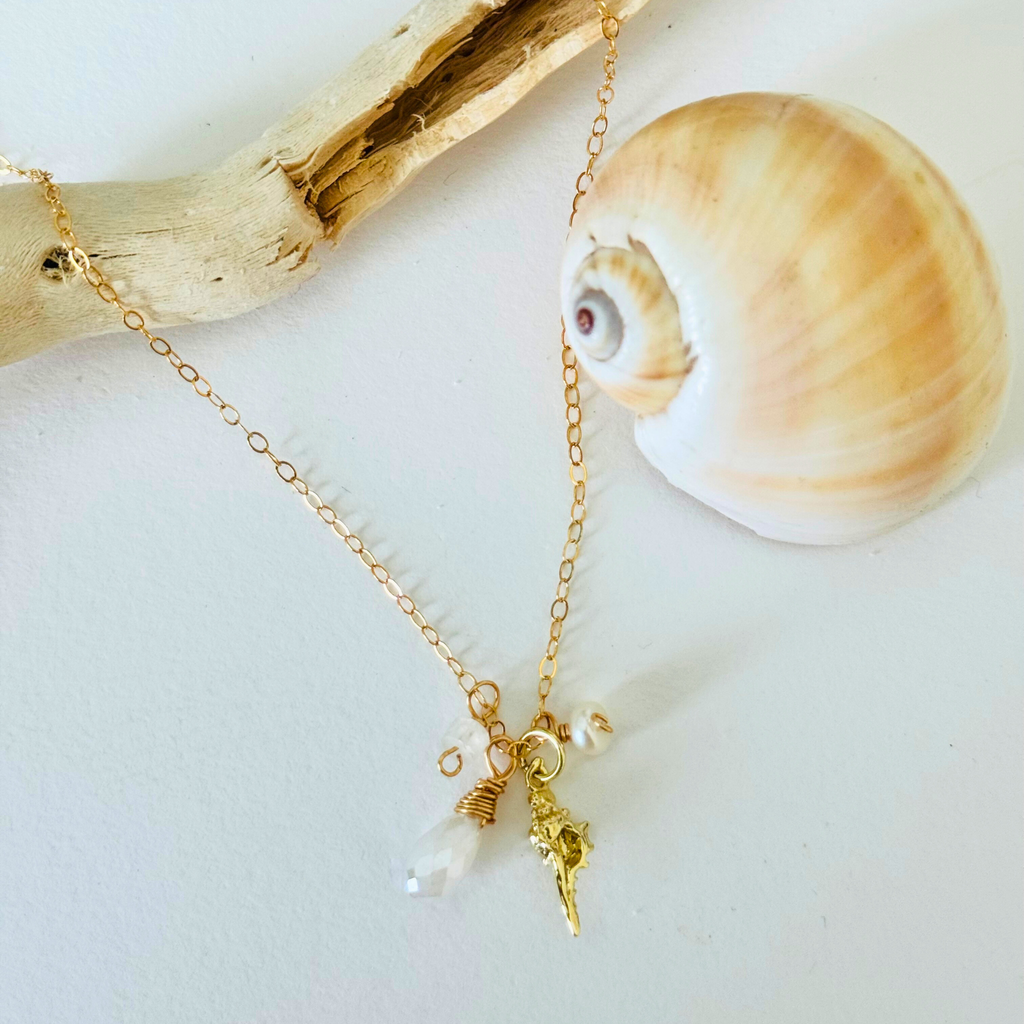 A simple dainty 14k gold filled wire necklace with pearl, aquamarine, and moonstone stones and a gold seashell charm are draped over bleached driftwood and framed by a creamy warm spiral seashell on a white background.