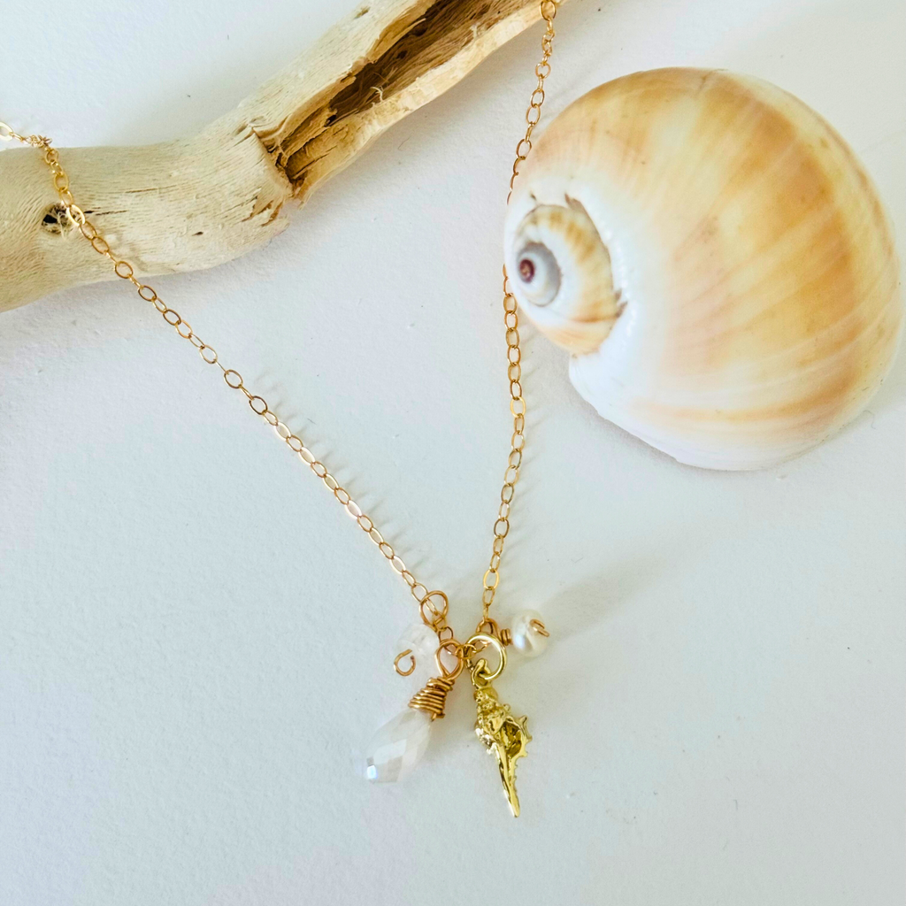 A simple dainty 14k gold filled wire necklace with pearl, aquamarine, and moonstone stones and a gold seashell charm are draped over bleached driftwood and framed by a creamy warm spiral seashell on a white background.