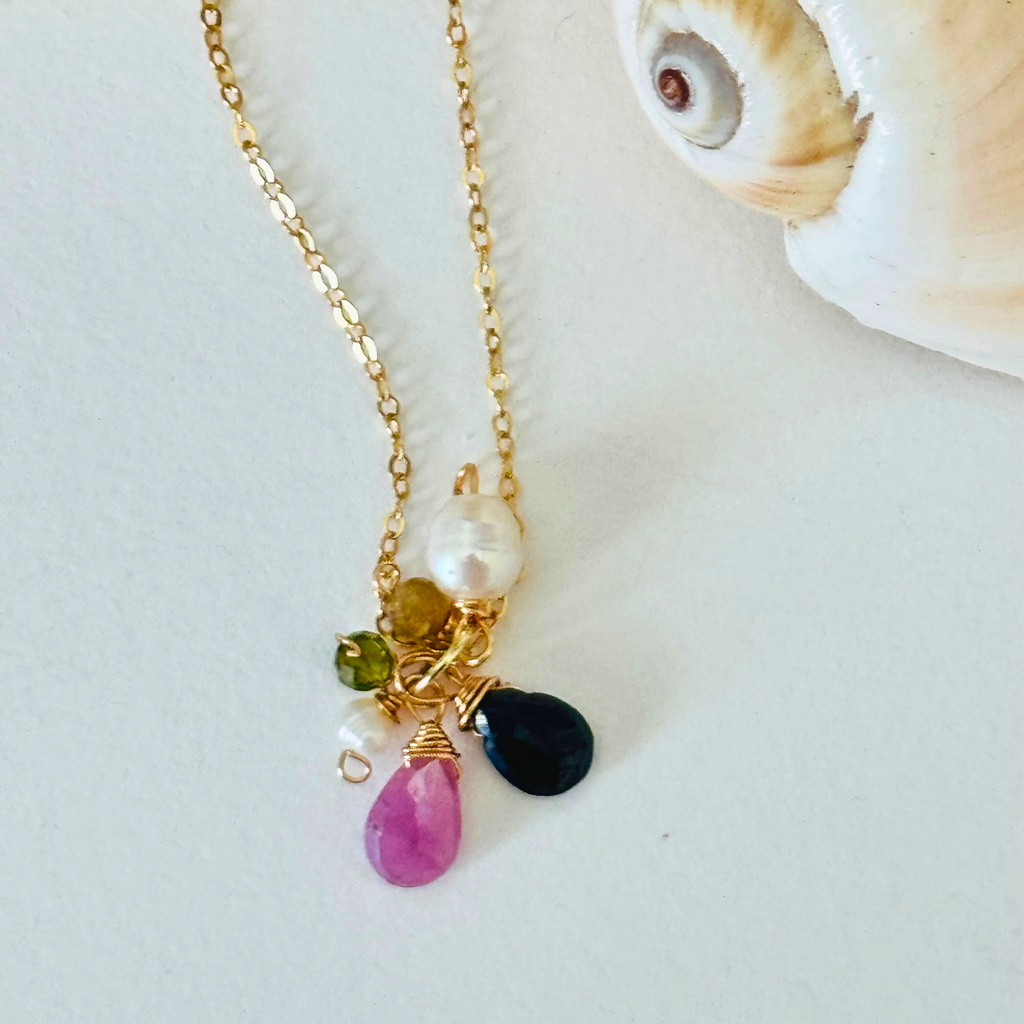 Closeup of pink sapphire, dark blue apatite, peridot, and freshwater pearl stones at end of handcrafted 14k gold filled wire necklace chain. Displayed on white background framed by a creamy spiral seashell.
