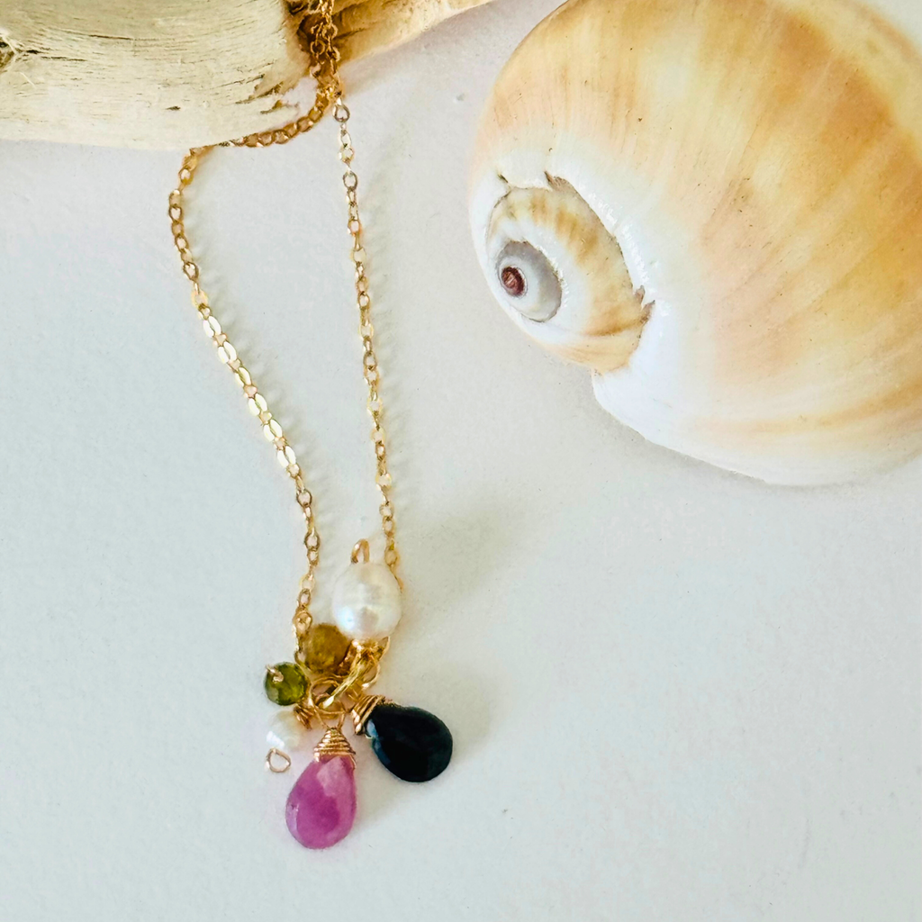 Closeup of pink sapphire, dark blue apatite, peridot, and freshwater pearl stones at end of handcrafted 14k gold filled wire necklace chain. Displayed draped over a bleached driftwood branch on white background framed by a creamy spiral seashell.