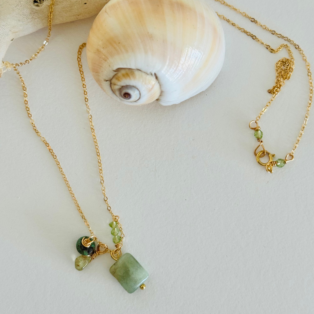  A necklace is draped over a creamy spiral seashell and bleached driftwood. It is made of a simple gold chain with aventurine, peridot, and turquoise. A pair of small light green beads offset the clasp.