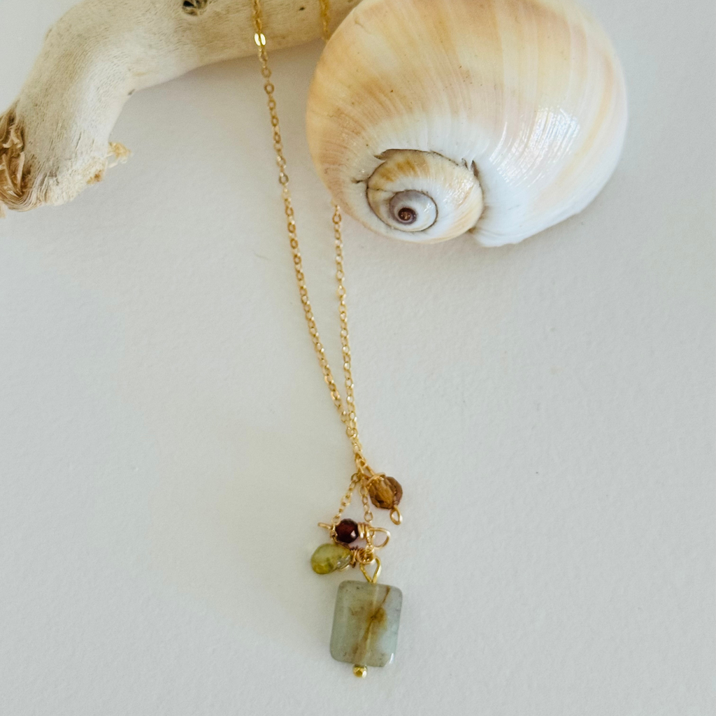 Simple chain necklace with one each of aventurine, peridot, and garnet hessonite beads in warm fall coastal colors displayed draped over a creamy spiral seashell and bleached driftwood.