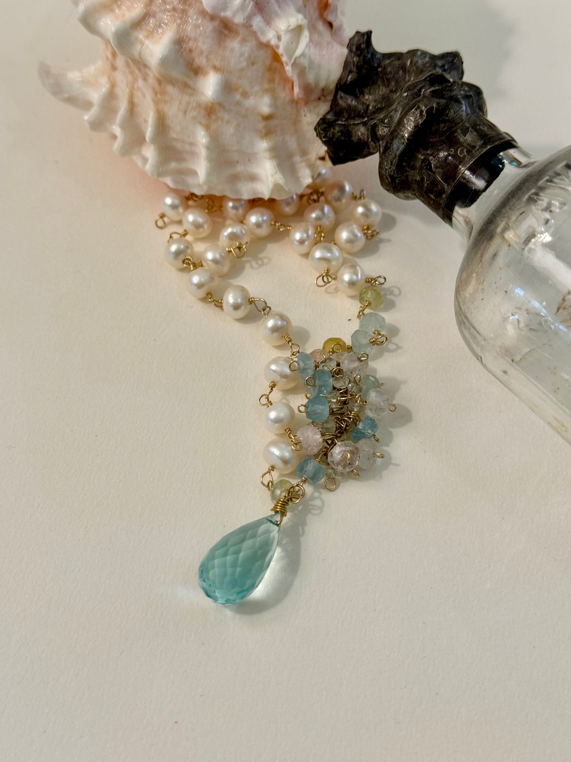 Pearl necklace with a blue teardrop pendant on a light surface with a shell and bottle.