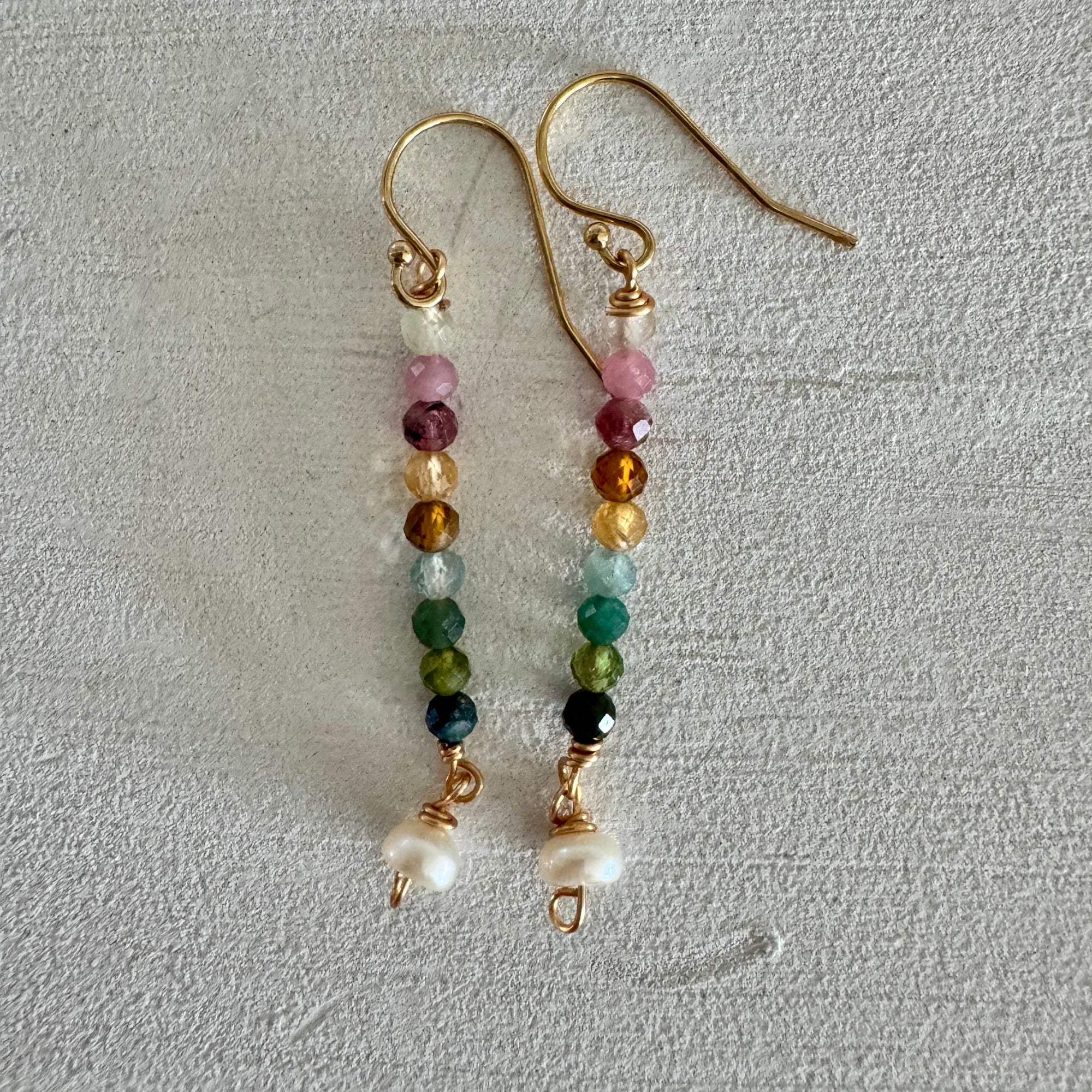 Multicolor beaded dangle earrings with pearl at the end on 14k gold filled wire displayed against a textured sand white background.