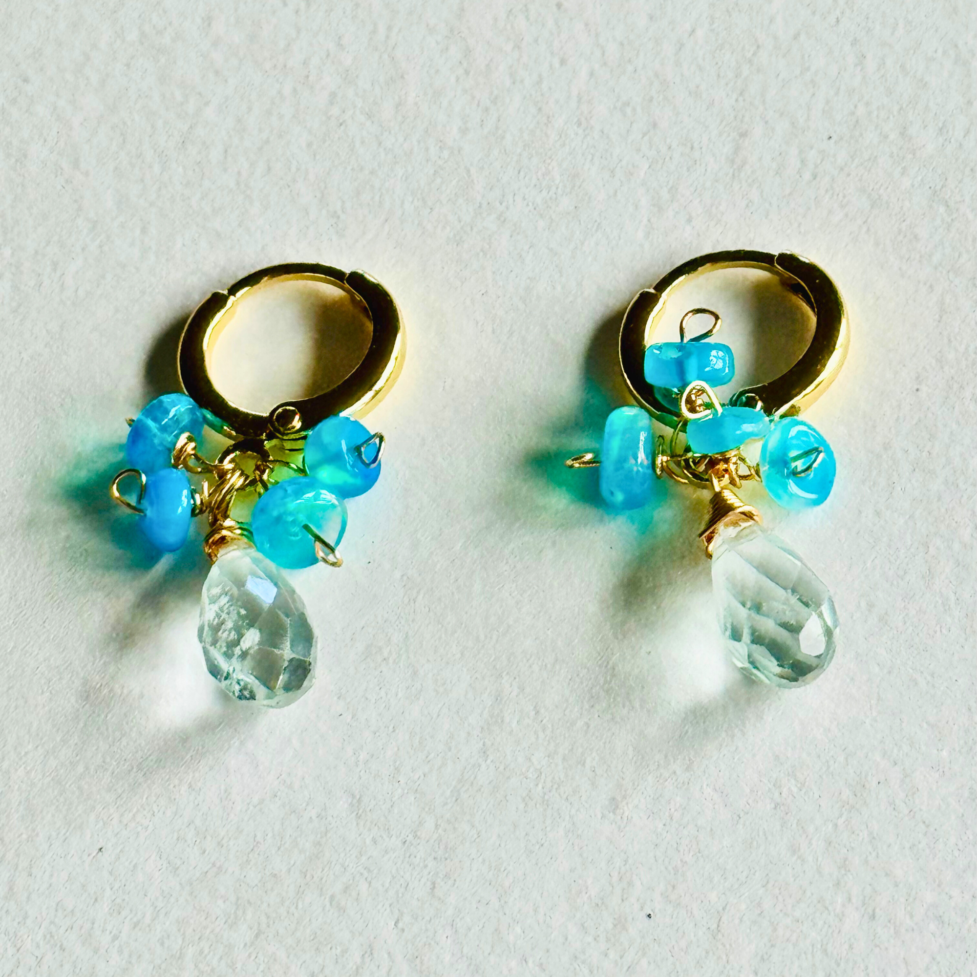 Pair of gold huggie earrings with blue beads and clear teardrop beads on a light background