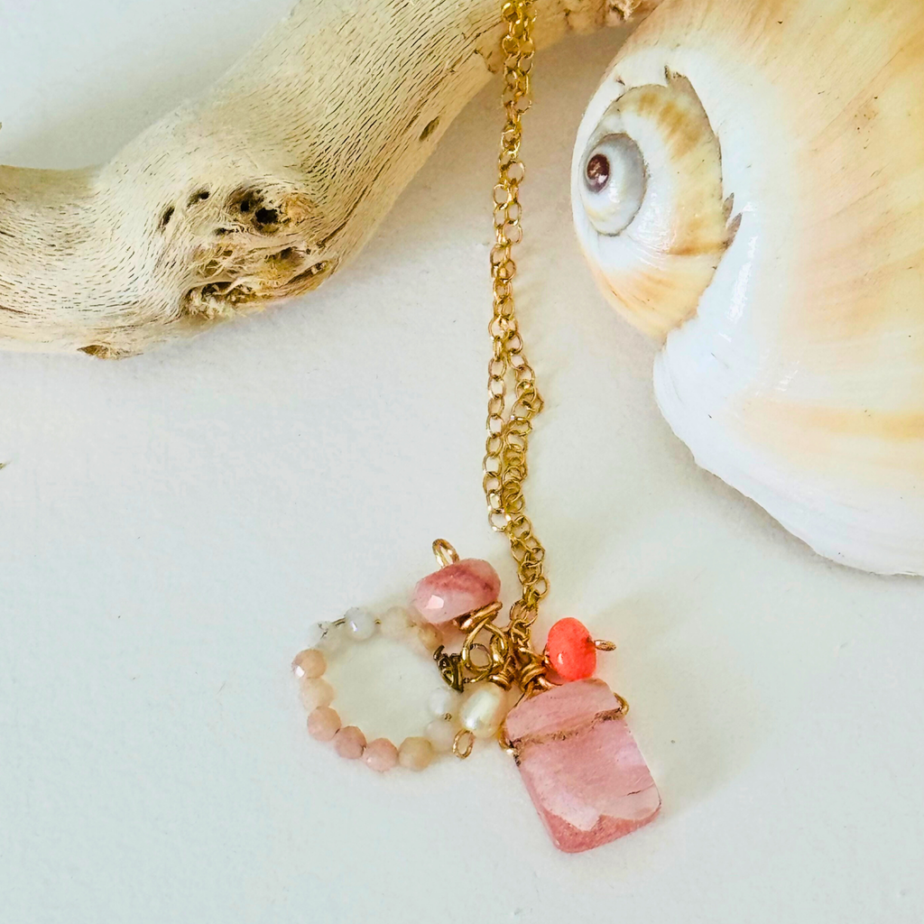 A dainty necklace made of rose quartz, pink Peruvian opal, and Ethiopian neon pink opal on handcrafted 14k gold filled wire chain is draped over bleached driftwood and framed by a creamy spiral seashell on a white background.