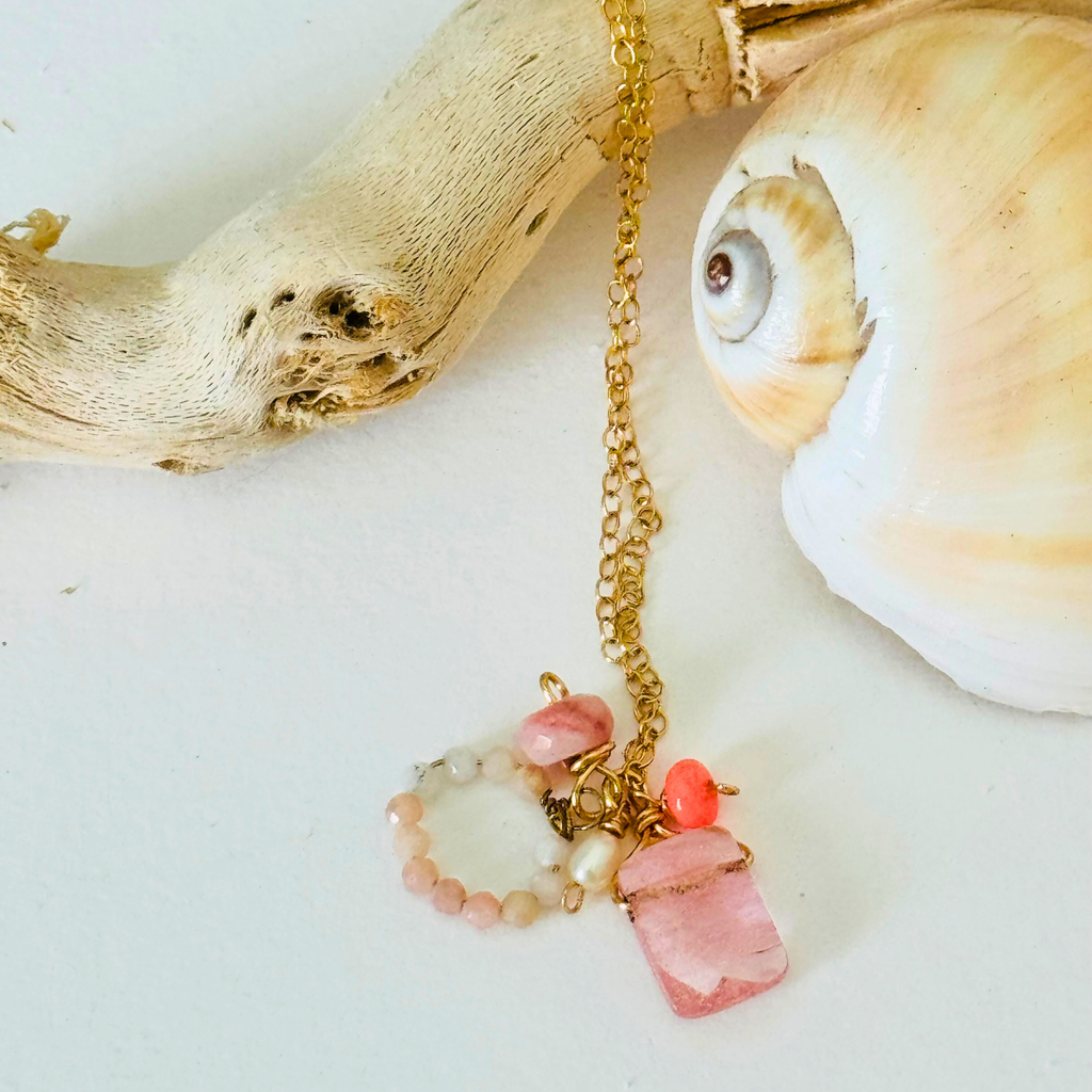 A dainty necklace made of rose quartz, pink Peruvian opal, and Ethiopian neon pink opal on handcrafted 14k gold filled wire chain is draped over bleached driftwood and framed by a creamy spiral seashell on a white background.