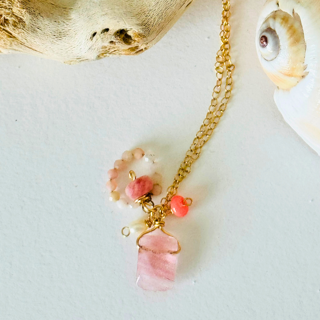 A dainty necklace made of rose quartz, pink Peruvian opal, and Ethiopian neon pink opal on handcrafted 14k gold filled wire chain is framed by bleached driftwood and a creamy spiral seashell on a white background.