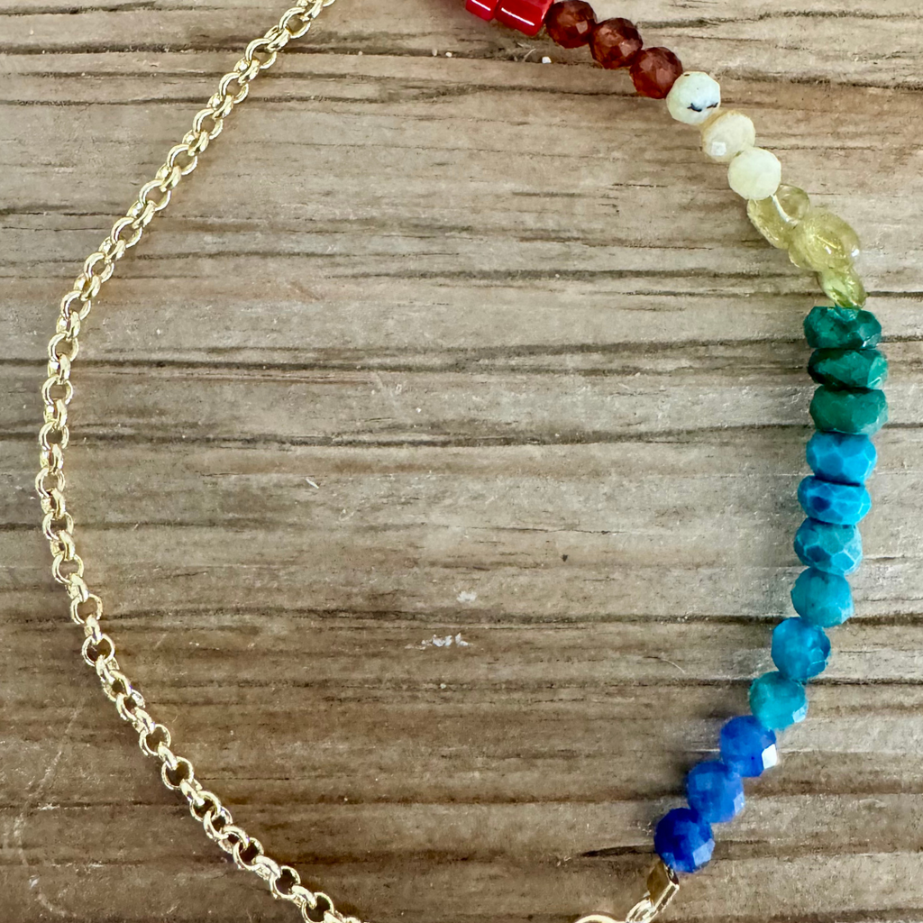 A bracelet is displayed on woodgrain surface. The bracelet is 14k gold chain with a pearl at the clasp. There are beads on half of the bracelet in three of each color of bright red, scarlet orange, milky yellow, glassy yellow-green, emerald green, blue, turquoise, and cerulean. Stunning rainbow ombre effect.