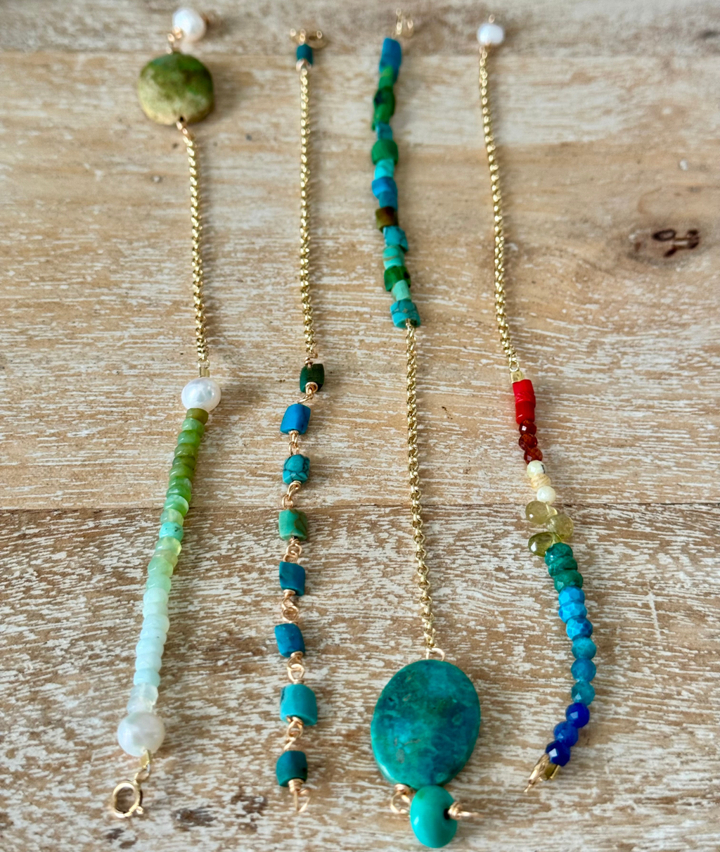 A set of four bracelets are laid out unclasped on a beachy wooden surface from 45-degree angle view. From left to right is a green seafoam opal pearl bracelet, blue and green square bead bracelet, turquoise opal bracelet, and beaded rainbow dreams bracelet, all with gold chains.