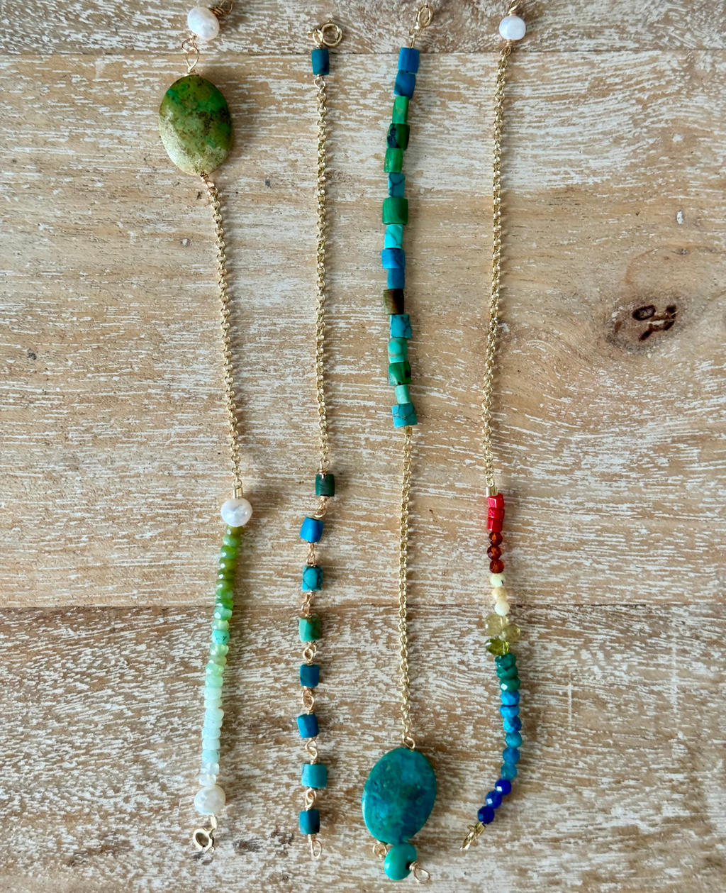 A set of four bracelets are laid out on a beachy wooden surface with an aerial view of them. From left to right is a green seafoam opal pearl bracelet, blue and green square bead bracelet, turquoise opal bracelet, and beaded rainbow dreams bracelet, all with gold chains.