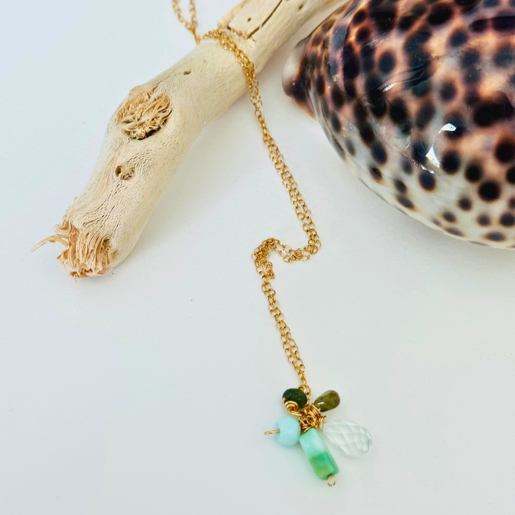 Closeup of amazonite, hessonite, and aquamarine stones at end of 14k gold filled wire chain on a white background draped over bleached driftwood and framed by a gleaming leopard-print shell.