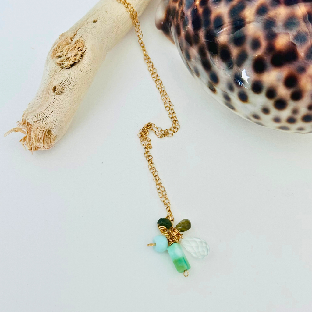 Closeup of amazonite, hessonite, and aquamarine stones at end of 14k gold filled wire chain on a white background draped over bleached driftwood and framed by a gleaming leopard-print shell.