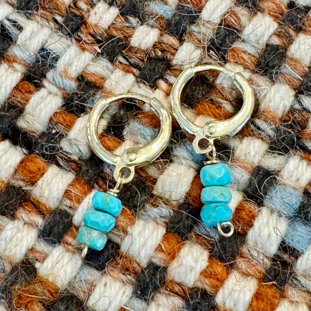 Close up view of gold-filled huggie earrings made with three wide round turquoise blue green stones stacked vertically, displayed against a white, orange, and brown textile background.