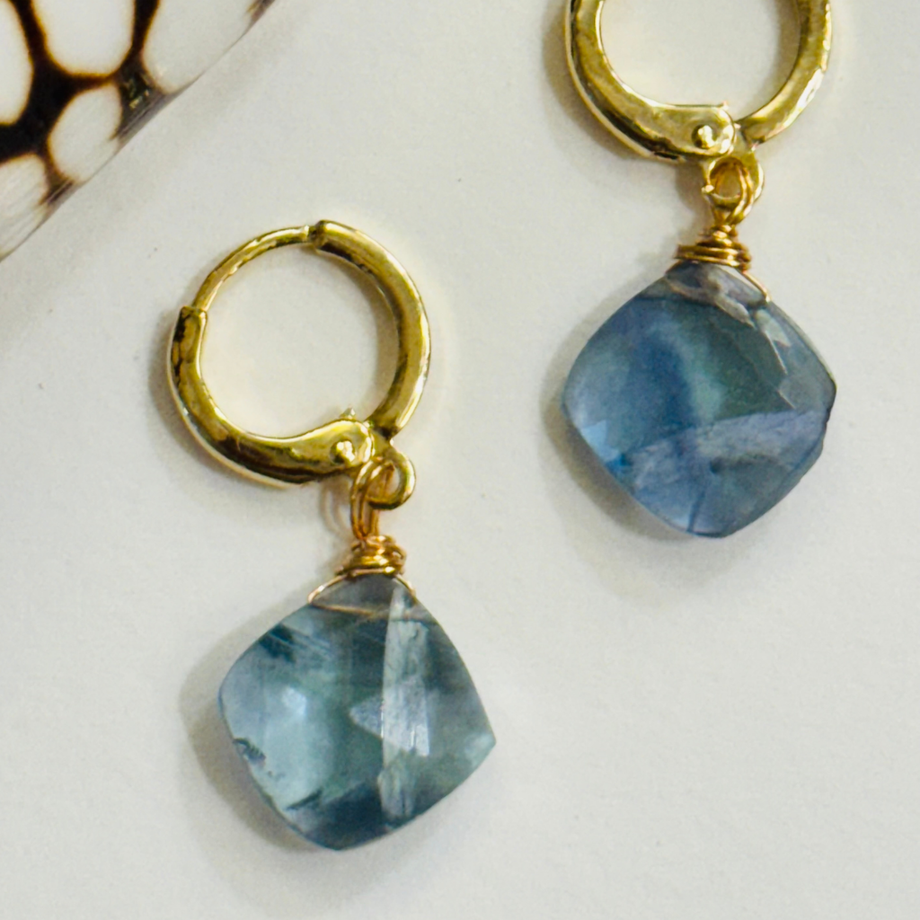 Closeup of a pair of earrings with a genuine teal fluorite gemstone on each 14k gold filled huggie hoop displayed on a white background.