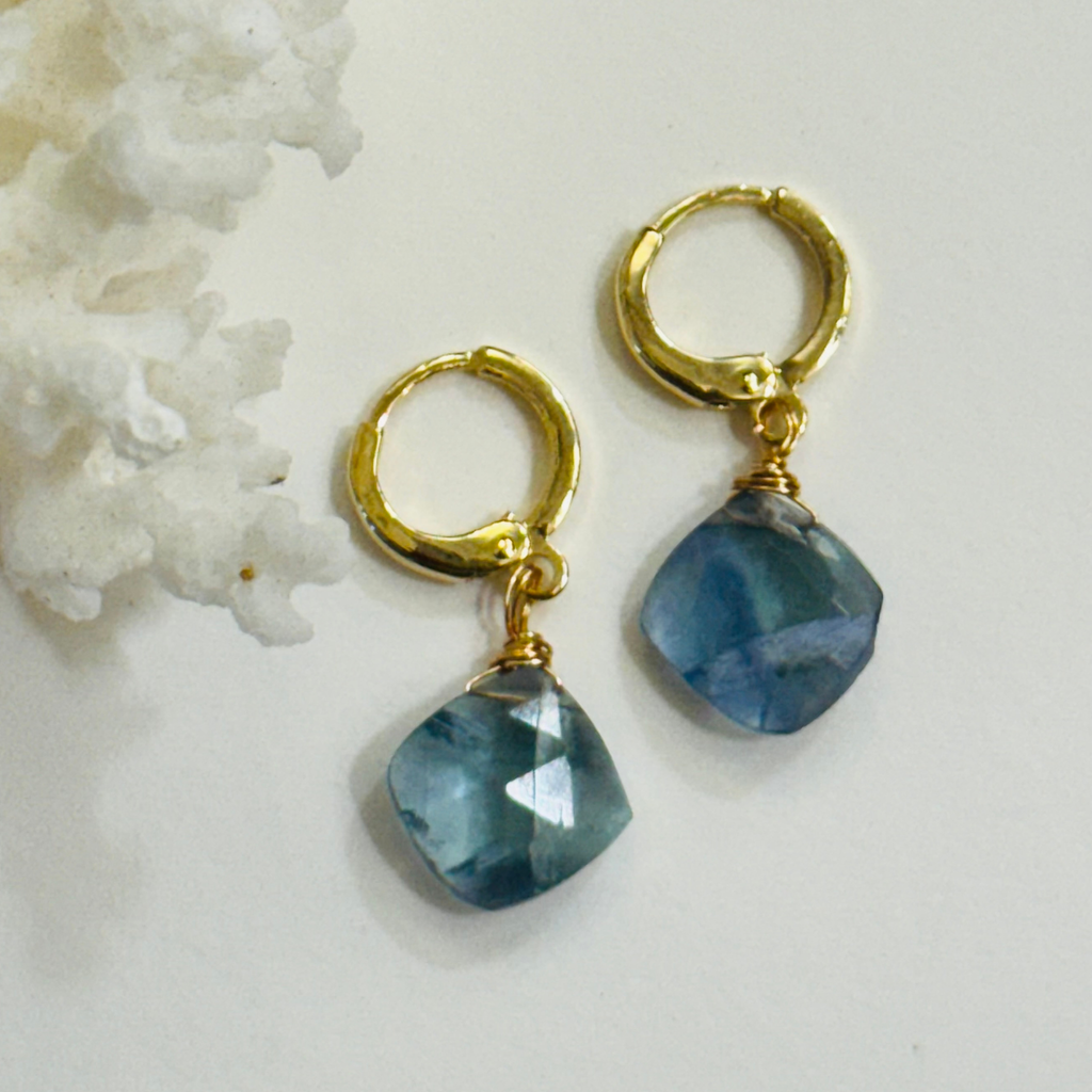 A pair of earrings with a genuine teal fluorite gemstone on each 14k gold filled huggie hoop is displayed on a white background and framed by white coral.
