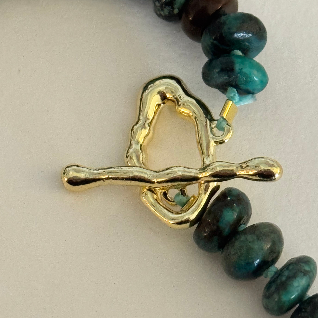 Closeup of wavy 14k gold filled toggle clasp and dark green and blue Hubei turquoise beads on a cream white background.