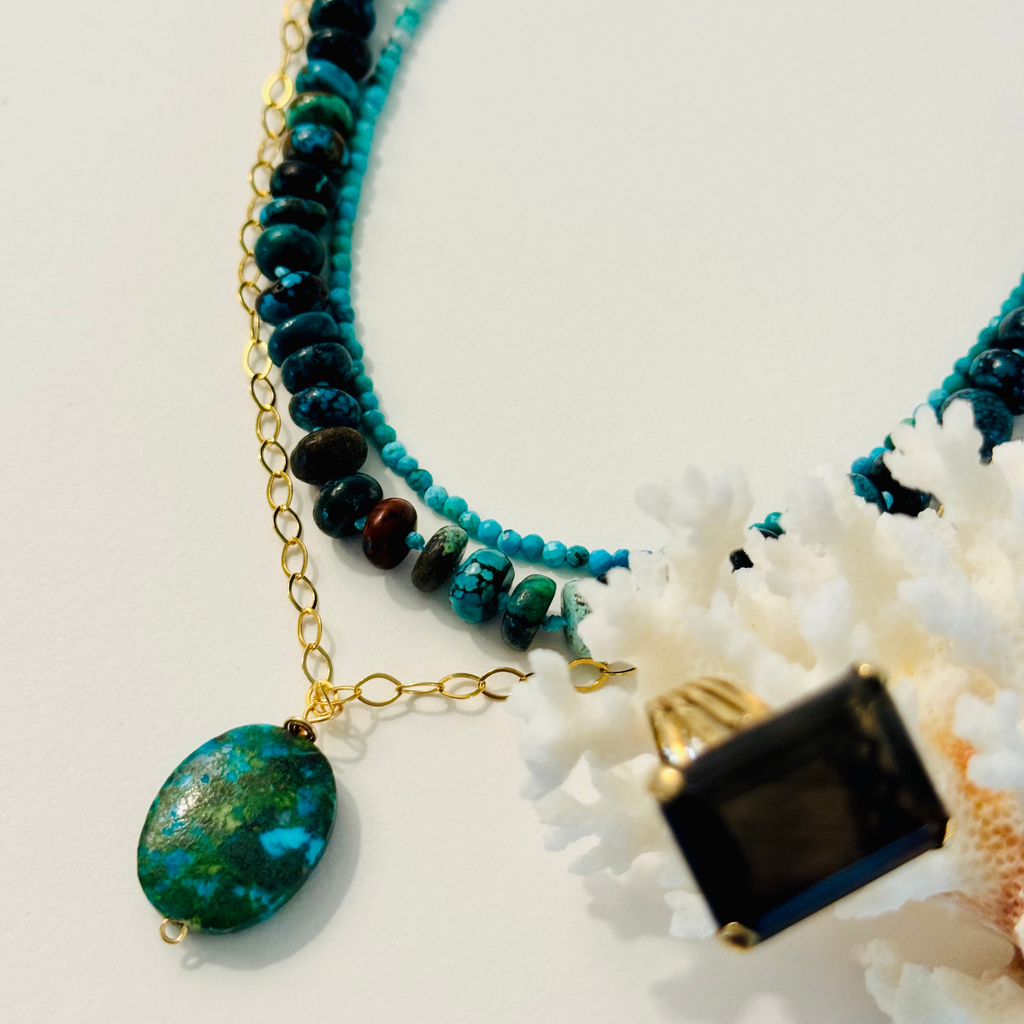 Layered stack of two beaded necklaces and one gold chain pendant necklace, all showcasing green and blue turquoise beads. Framed by white coral and navy and gold ring on a white background.