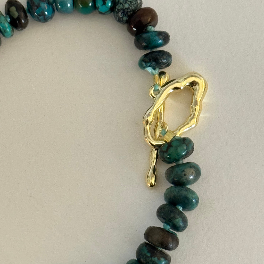 Genuine Hubei turquoise plain rondelle beads in varying shades of Cerulean blue, navy, green, and in between hand knotted on silk cord with a wavy 14k gold filled toggle clasp on a cream white background.