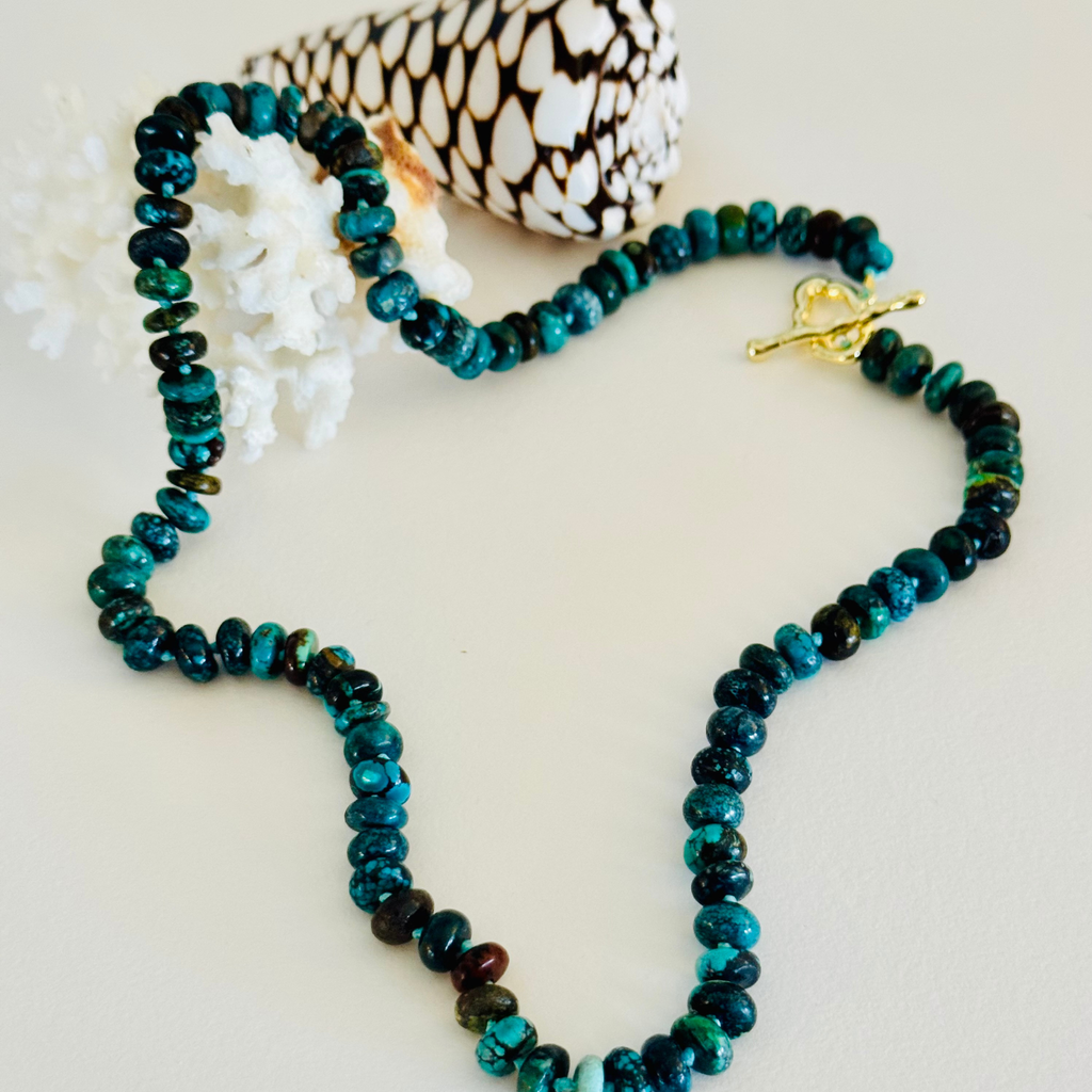 Genuine Hubei turquoise plain rondelle beads in varying shades of Cerulean blue, navy, green, and in between hand knotted on silk cord with a wavy 14k gold filled toggle clasp. Displayed draped over white coral, framed by a white and brown spotted cone spiral seashell on a cream white background.