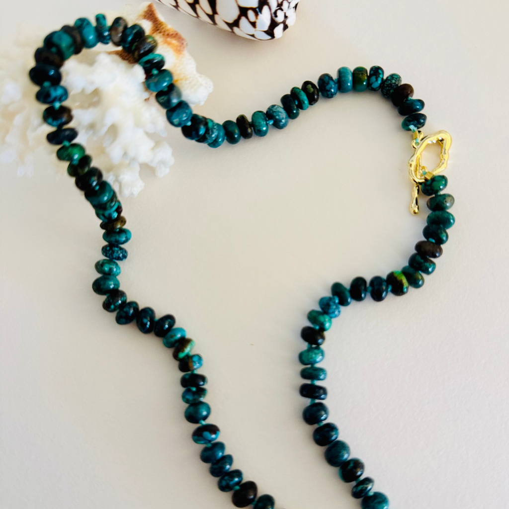 Genuine Hubei turquoise plain rondelle beads in varying shades of Cerulean blue, navy, green, and in between hand knotted on silk cord with a wavy 14k gold filled toggle clasp. Displayed draped over white coral, framed by a white and brown spotted cone spiral seashell on a cream white background.